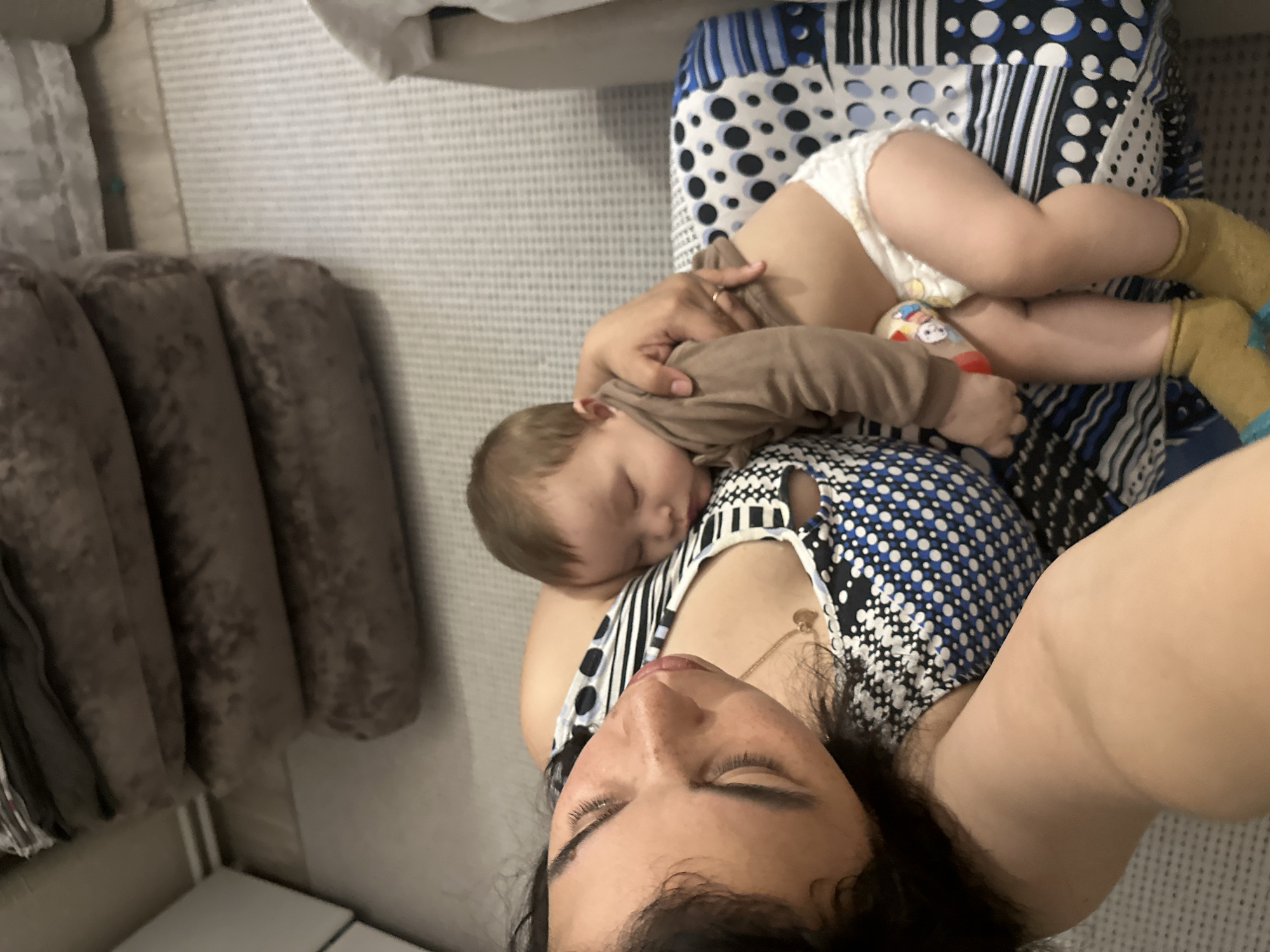 This image captures a serene indoor scene depicting a woman and a baby, likely a mother and her child, resting together. The photograph is oriented vertically, showing the subjects lying on their side from a slightly elevated perspective.
The woman is in the foreground, with her head and upper body visible. She has dark hair and her eyes are closed, suggesting she is asleep or in a state of deep relaxation. She is wearing a sleeveless top with a distinctive pattern of black, white, and blue, incorporating both stripes and polka dots. A thin gold chain with a small charm is visible around her neck.
Cradled on the woman's chest or shoulder is a baby, also appearing to be fast asleep. The baby has light brown hair and is dressed in a plain, light brown long-sleeved top. A white disposable diaper is visible, and the baby's feet are covered in yellow or light brown socks. A small, colorful toy, possibly a rattle or soft character, is partially visible near the baby's hand, resting on the diaper.
The background reveals a comfortable, domestic setting. Beneath the mother and baby is a patterned fabric, likely part of a blanket, bed, or armchair. To the left, a plush, brown upholstered surface, resembling cushions or a headboard, provides a soft backdrop. The floor beneath them appears to be covered with a light-colored, textured carpet or rug.
The image exudes a sense of peace and intimacy, capturing a quiet moment of rest and bonding between the mother and child. There is no visible text in the image.