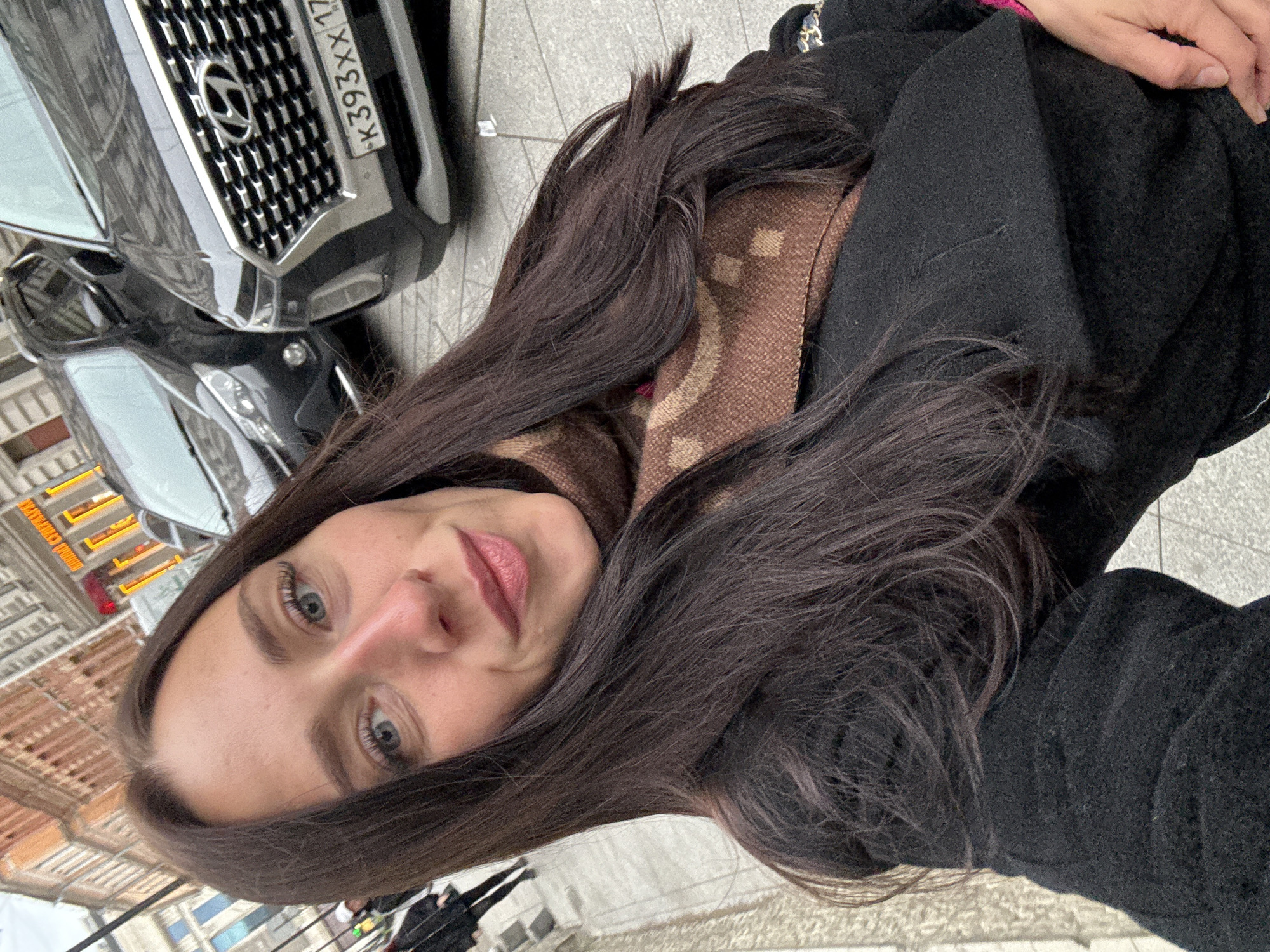 A woman takes a selfie outside, near a black Hyundai car with the license plate "K393XX 177" indicating its registration in Moscow. Based on architectural details like the buildings and a street sign visible in the background, the location is likely Saint Petersburg, Russia. She wears a black coat and a brown patterned scarf. Other pedestrians are visible in the background. The ground appears to be paved.