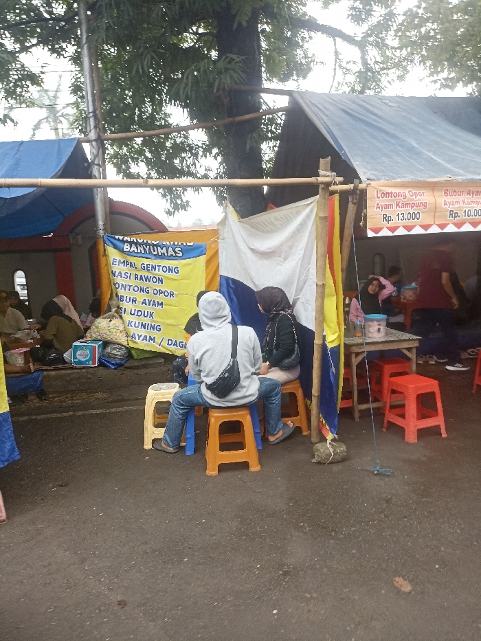 The image shows a street food vendor in Purwokerto, Indonesia, located at coordinates -7.417, 109.249. The vendor is set up under a blue tarp supported by a bamboo frame. A large sign advertising "Banyumas" food items is visible. Two people are sitting on yellow stools, facing away from the camera, while other individuals appear to be waiting for their food. A white plastic stool is partially visible on the right. A blue and yellow flag is in the background. On the far right, a table and chairs are visible. A stone is in the foreground. There is a sign advertising "Lontong Opor Ayam Kampung" for Rp. 13,000 and a sign advertising "Bubur Ayam Kampung" for Rp. 10,000. There are multiple people in the scene, but only the backs of the two people on the yellow stools are visible. The overall environment is that of a busy street food stall in a busy city.