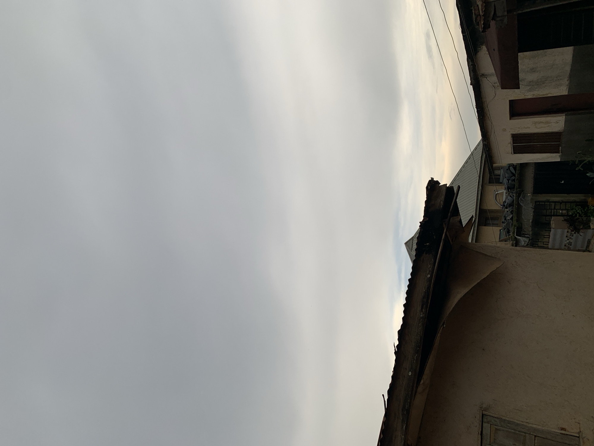 The image captures a street scene in Rafin Par, Nigeria, under an overcast sky. In the foreground, the side of a building constructed with rough, light brown plaster is visible. A dilapidated wooden window with a weathered frame is set into the wall. Above this building, the edge of a corrugated metal roof, darkened with age and possibly soot, is prominent. Further into the scene, other buildings with pitched roofs are visible. One building to the left has a grey, ribbed metal roof. Another building to the right features a flat roof with a light-colored facade and a dark red door. Overhead, electrical wires are strung between poles. The overall impression is one of a densely built urban environment, with signs of wear and tear on the structures. There are no people or animals visible in the frame.