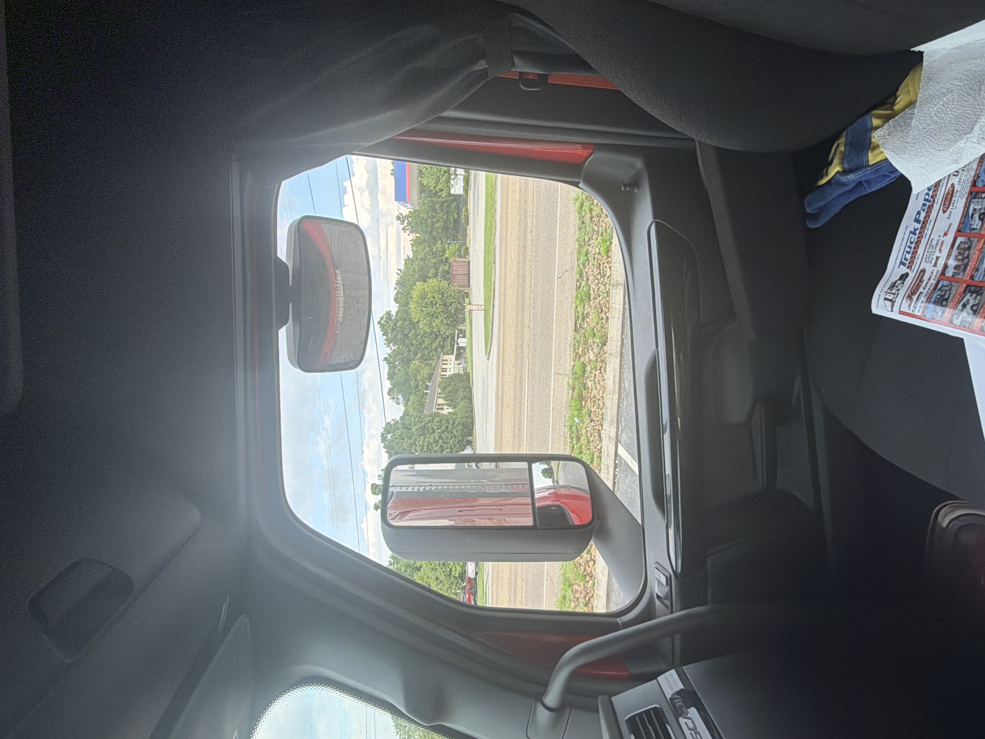 The image is taken from inside a truck cab, looking out the driver's side window. The truck appears to be stopped, possibly at an intersection or parking lot. Outside the window, a road is visible, with a sidewalk and a grassy area. Beyond that, there are trees, buildings, and a gas station with a red and blue sign. In the foreground, inside the truck, a newspaper titled "Truck Paper" is visible on the passenger seat, along with a pair of work gloves. The sky is cloudy. No people are visible in the image. The location is identified as Chattanooga, United States of America.