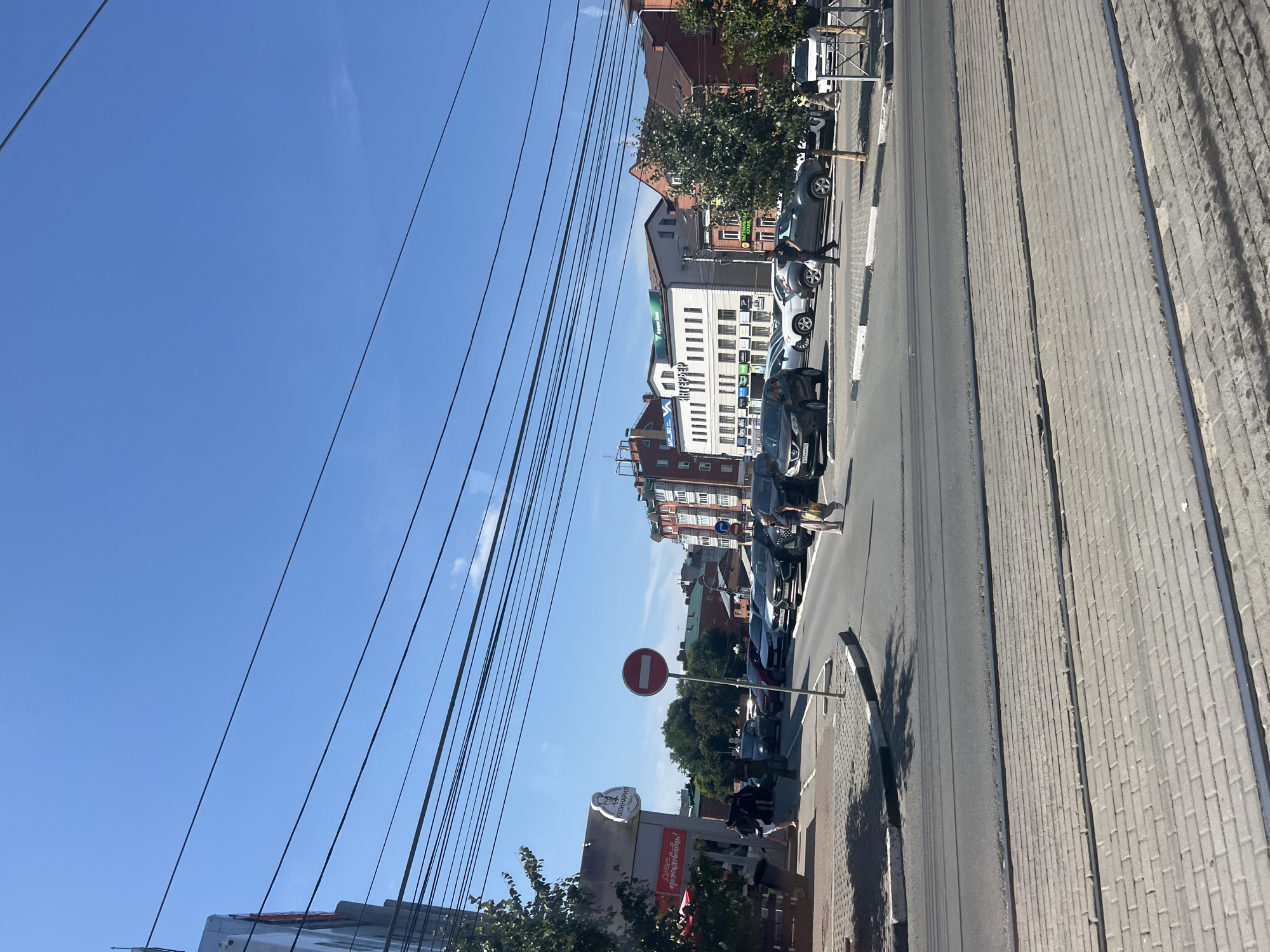 The image depicts a street scene in the city of Ulyanovsk, Russia, on a clear, sunny day. Several cars are parked along the street and a few are in motion. People are walking on the sidewalk and crossing the street. Overhead, a network of power lines crisscrosses the frame. The buildings lining the street appear to be a mix of residential and commercial properties, with signage visible in Russian. A "no entry" sign is prominently displayed near an intersection. The foreground shows tram tracks embedded in a brick-paved walkway.
