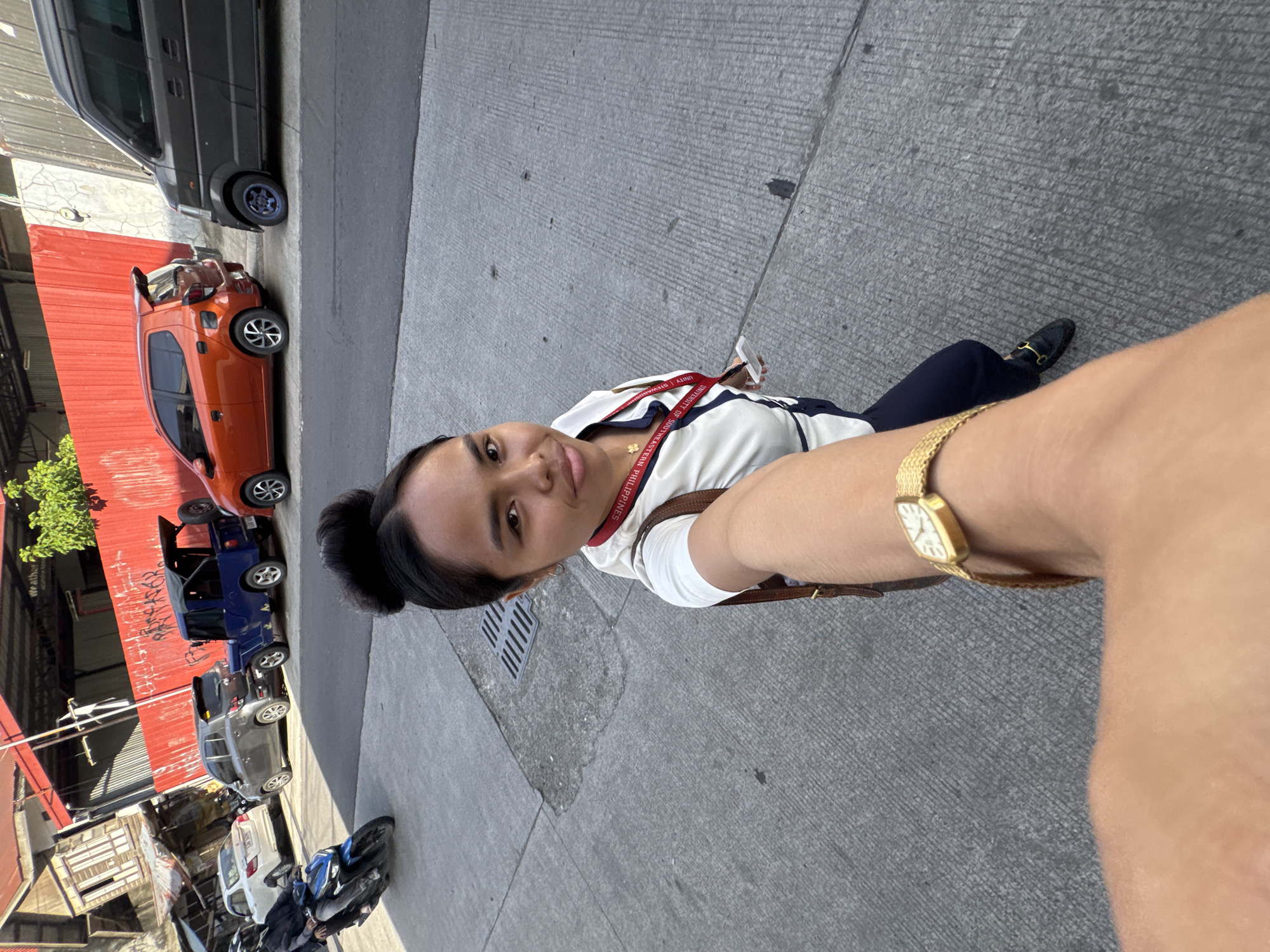 A young woman with her hair in a bun, wearing a white and navy uniform with a lanyard, stands on a concrete sidewalk, taking a selfie with her right arm extended. She wears a gold wristwatch on her left wrist. She is dressed in dark trousers and black loafers with gold accents. In the background, several vehicles are parked along a street, including an orange hatchback, a blue open-top vehicle, a gray SUV, a white sedan, and a dark gray van. A motorcycle with two riders is also visible on the road. The street is lined with buildings, one of which has a bright red facade. The overall setting suggests an urban environment in Davao City, Philippines, given the text on her lanyard which reads "UNIVERSITY OF SOUTHEASTERN PHILIPPINES". The lighting indicates it is daytime.