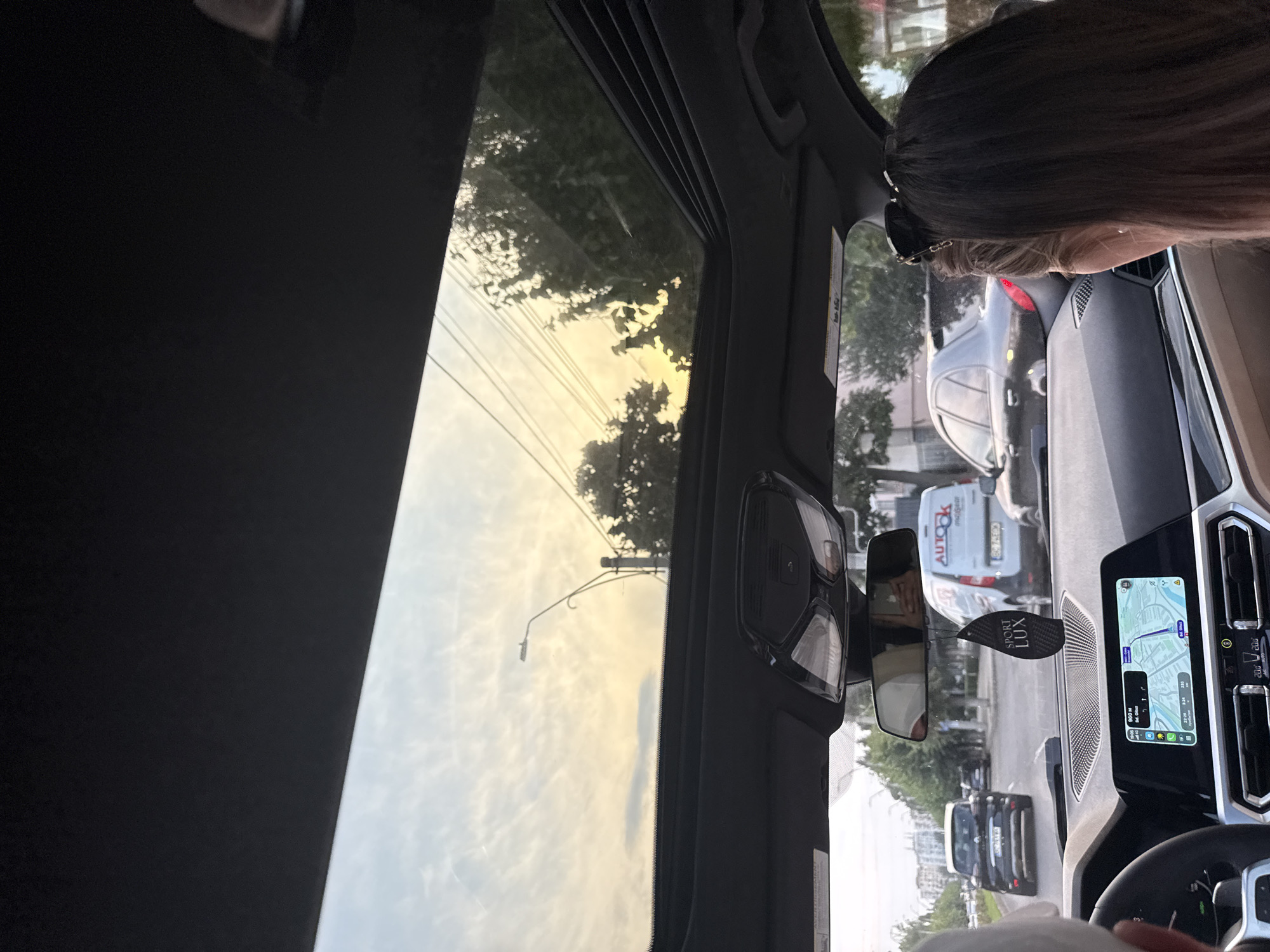The image shows the interior of a car in motion, looking out through the sunroof and windshield. The scene appears to be a street in the city of Onești, Romania, as indicated by the presence of a van with "AUTOK" branding, a common sight in Romanian businesses. The time of day is late afternoon or early evening, with a warm, golden sunset visible through the sunroof, casting a soft light on the clouds.
In the foreground, the dashboard of the car is visible, with a navigation system displaying a map. The steering wheel and parts of the car's interior are also present, suggesting the viewpoint is from the driver's or a passenger's seat. A woman with dark hair and sunglasses perched on her head is visible on the right side of the frame, looking out the side window.
Outside the car, traffic is visible on a city street. A black SUV is in front of the car, followed by other vehicles. A white van with "AUTOK" and "SPORT LUX" branding is also present in the traffic. Trees and buildings line the street, typical of an urban environment. The overall impression is of a mundane moment of travel through a Romanian city at dusk.