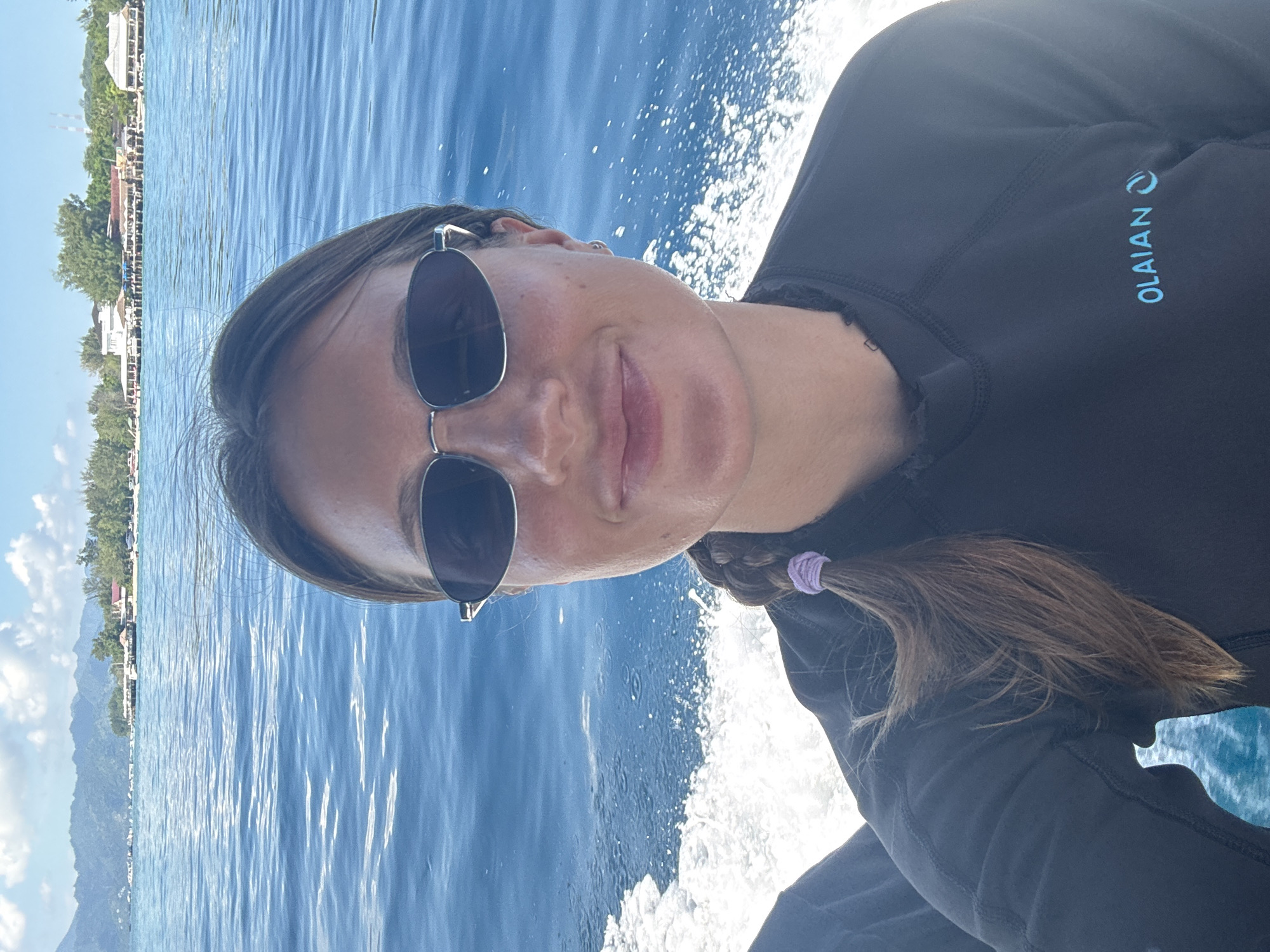 A young woman is posing for a selfie while on a boat. She is wearing sunglasses and a black wetsuit with the word "OLAIAN" on the sleeve. Her hair is pulled back in a braid with a purple hair tie. The background shows a body of water with a coastline featuring buildings, trees, and what appears to be a mountain range. The location is likely in Unknown, Indonesia.