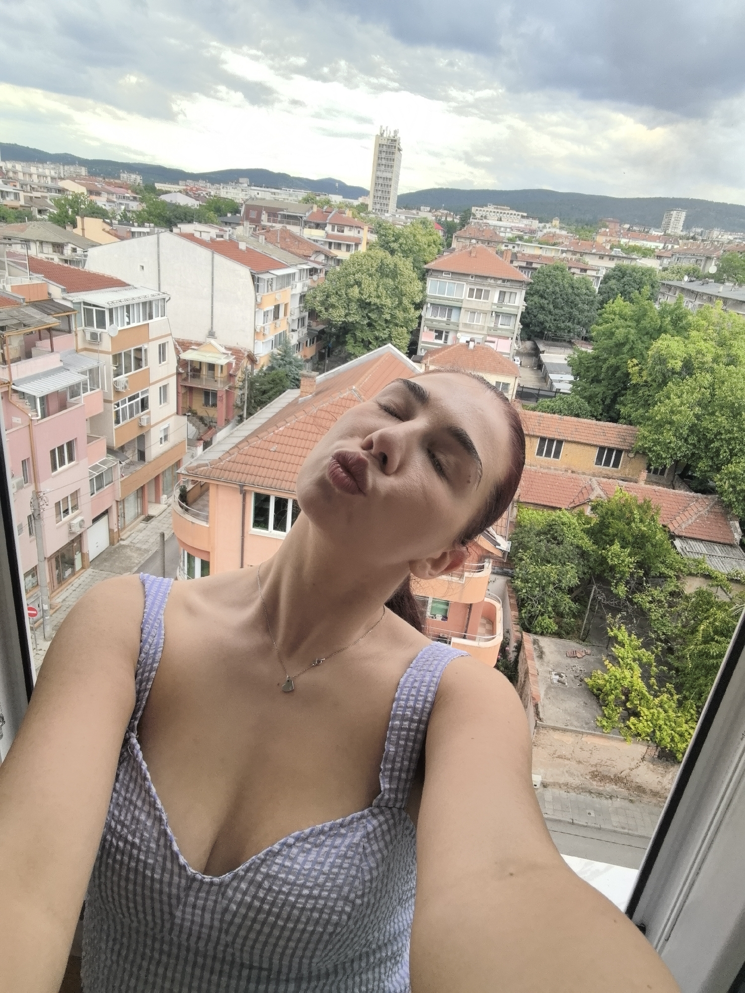 A woman is taking a selfie by an open window. She is looking towards the camera with a pouting expression. She has dark hair pulled back in a ponytail and is wearing a blue and white striped top. A necklace with a heart-shaped pendant is visible on her chest.
Outside the window, there is a cityscape of Stara Zagora, Bulgaria. Buildings of various heights and colors with red-tiled roofs dominate the view. A tall white tower is seen in the distance. Trees add green patches in between the buildings. The sky is overcast with grey clouds.