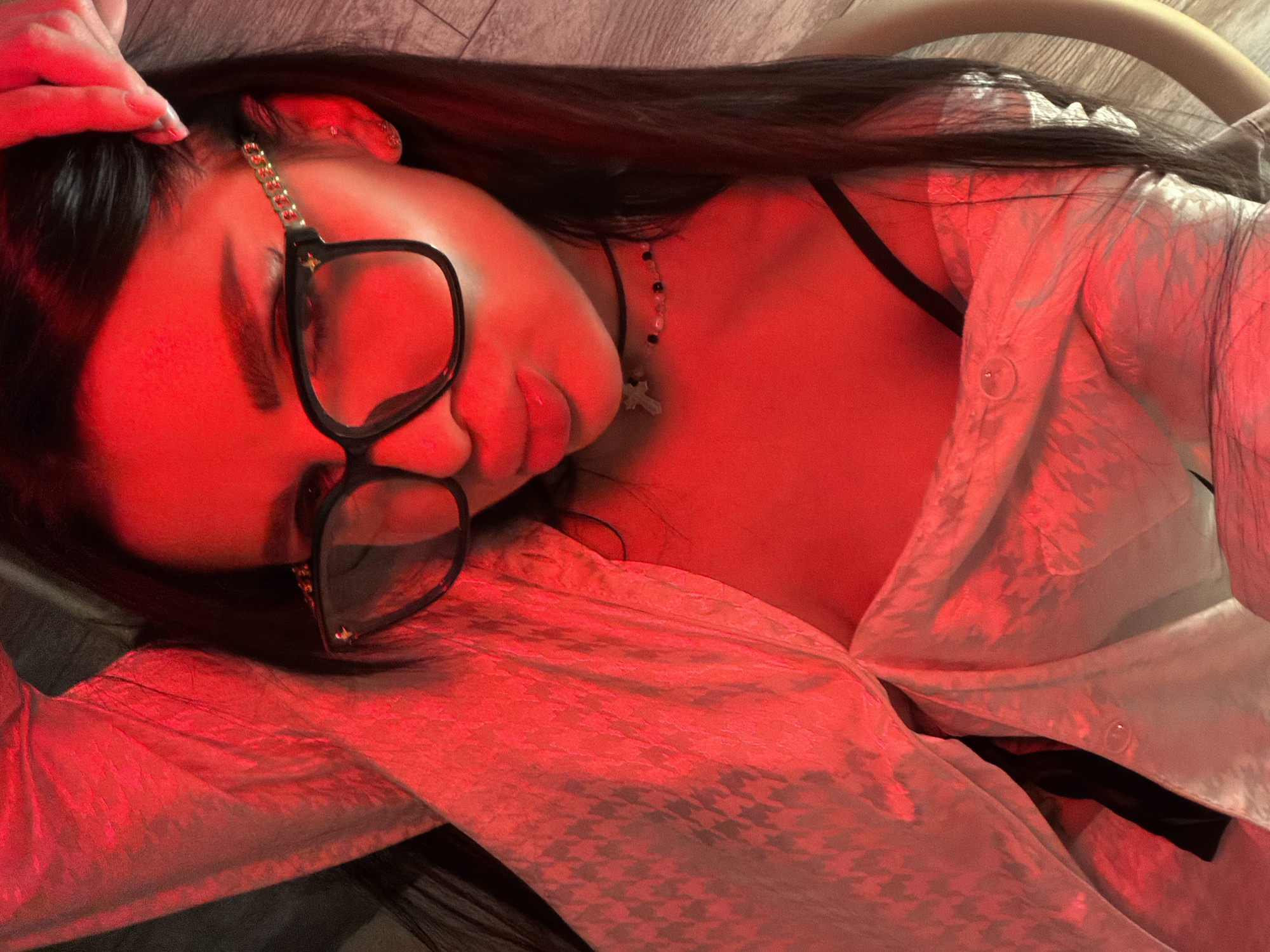 A person lies on their back in an indoor setting. They are wearing glasses with a thick black frame and a gold chain detail. A necklace with a cross pendant is visible around their neck. They are also wearing a white button-down shirt, revealing a black bra underneath. The scene is lit with a strong red light. Moscow, Russia is not depicted in the image.