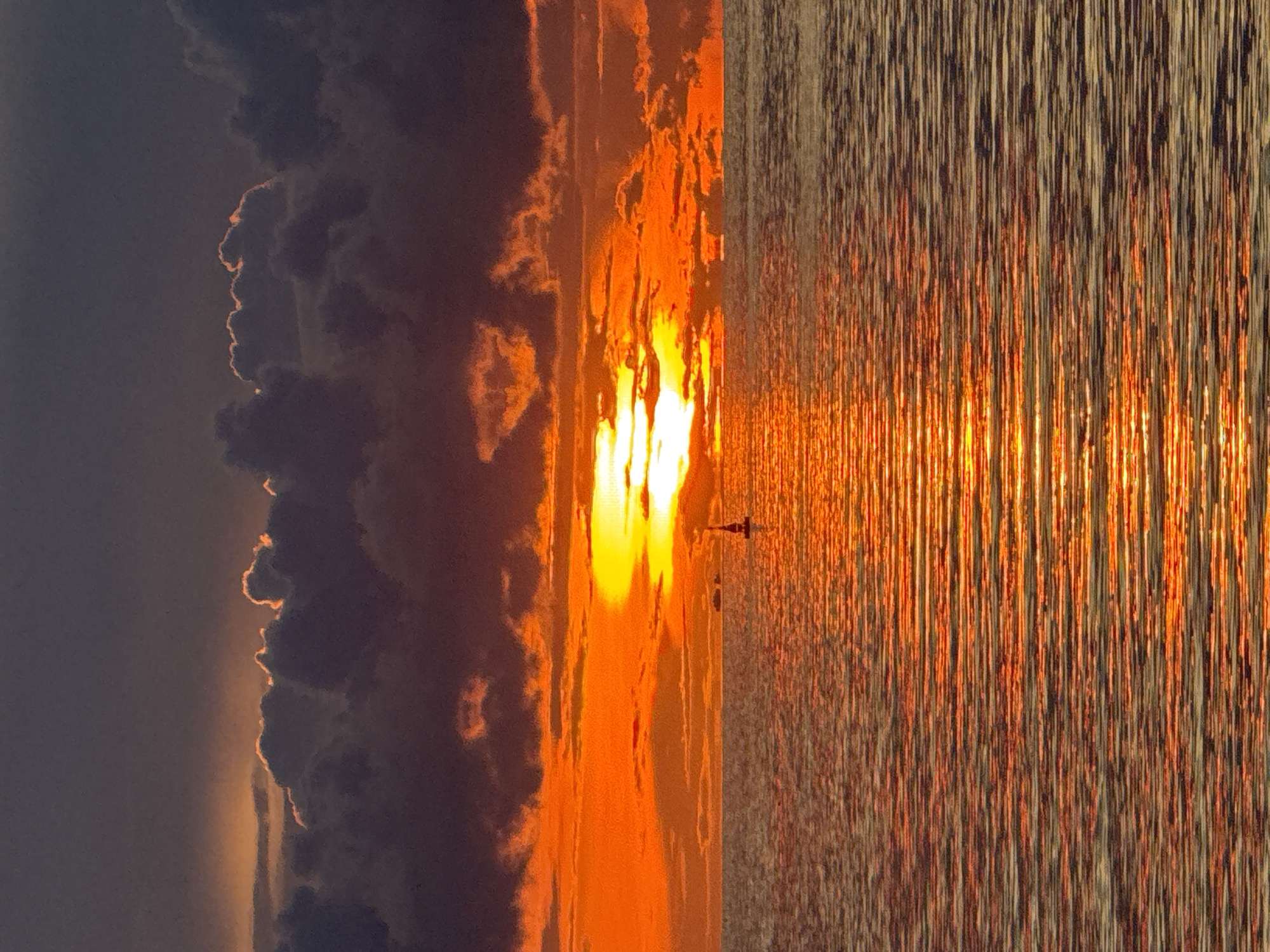 The image captures a sunset over water, likely the sea or a large body of water, in Baan Wok Tum, Thailand. The sun, a brilliant, intense white, is positioned centrally in the frame, with a vertical orange reflection radiating across the water's surface. Dark, textured clouds occupy the left side of the image, the edges of which are illuminated by a soft, orange glow from the setting sun. The water displays an intricate pattern of vertical lines, indicating wave movement and light reflection. There is an indication of an object in the water, possibly a boat.