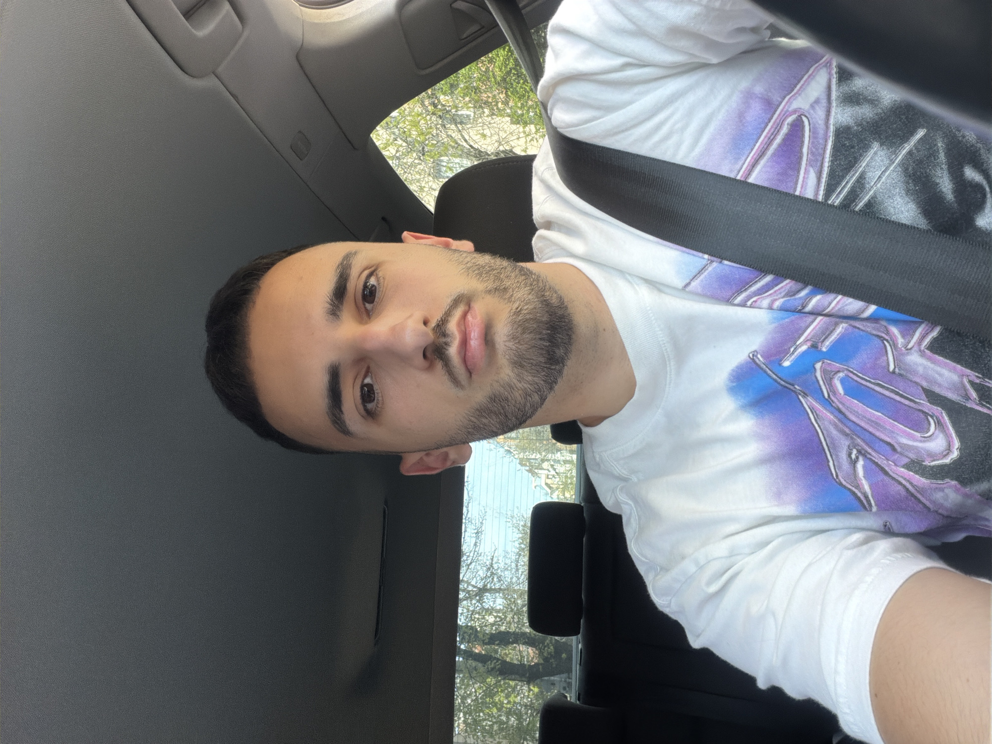 A man is seated in the driver's seat of a car, wearing a white t-shirt with a graphic design and a seatbelt. Outside the car window, trees are visible. The location cannot be determined from the image. There are no identifiable landmarks indicating the city of Plovdiv, Bulgaria.