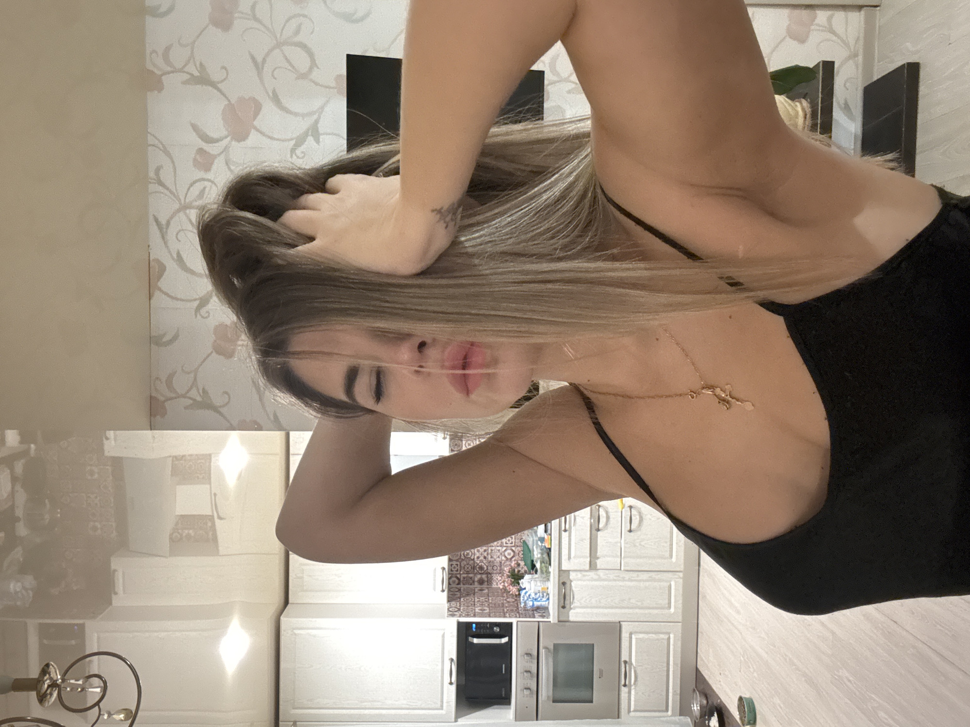 A young adult female with light brown hair, closed eyes, and slightly parted lips is depicted indoors, presumably located in Chaikovsky, Russia. She wears a black tank top and a gold necklace with a crucifix pendant. Her arms are raised, and her hands are positioned within her hair. A small, stylized tattoo is visible on her left wrist.
The background reveals a residential interior. To the left, a modern kitchen area features white cabinetry with silver handles, an integrated stainless steel microwave above a matching oven, and a tiled backsplash with brown and white mosaic patterns. Various items, including what appears to be a floral arrangement and containers, are visible on the kitchen counter. The floor in this area is light-colored wood laminate. To the right, a wall is covered with wallpaper featuring a light background with a repeating floral and vine pattern in pink and brown tones. A black, rectangular object, possibly a television or monitor, is mounted on this wall, with two dark-colored wall shelves beneath it. The ceiling is a reflective white surface, showing reflections of an ornate chandelier visible in the upper left corner and other light sources.