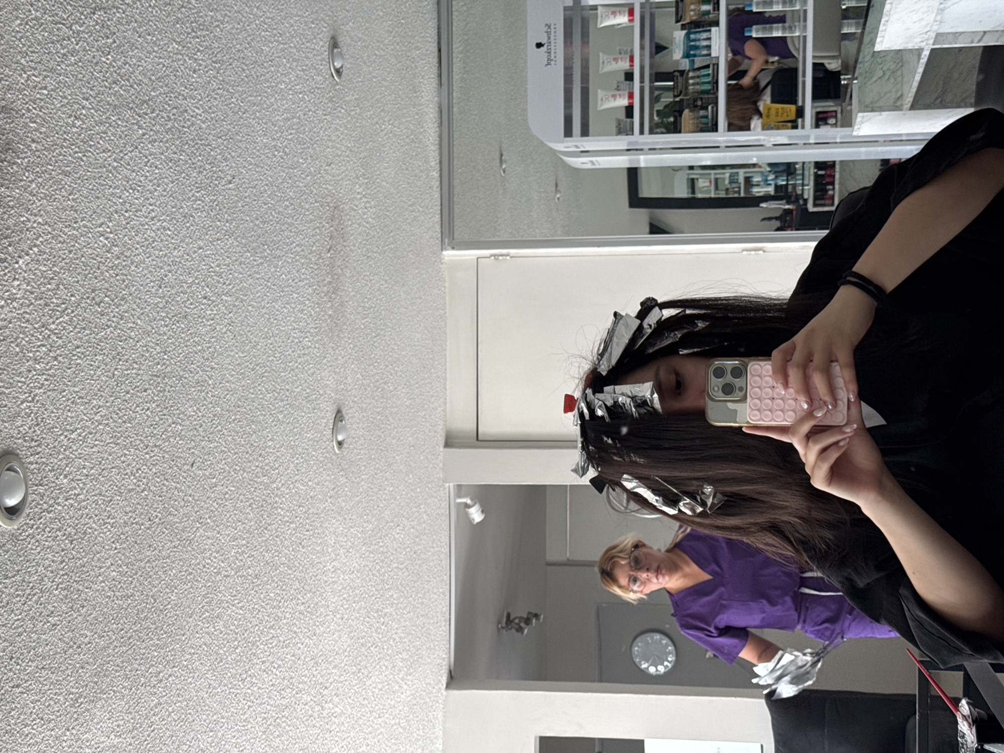 The image depicts a person taking a selfie in a hair salon, with foil highlights in their hair. The reflection shows a stylist in a purple uniform. In the background, there is a shelf stocked with hair care products and a clock on the wall. The location appears to be a salon interior. There is no confirmed evidence for this image being in Puebla, Mexico.