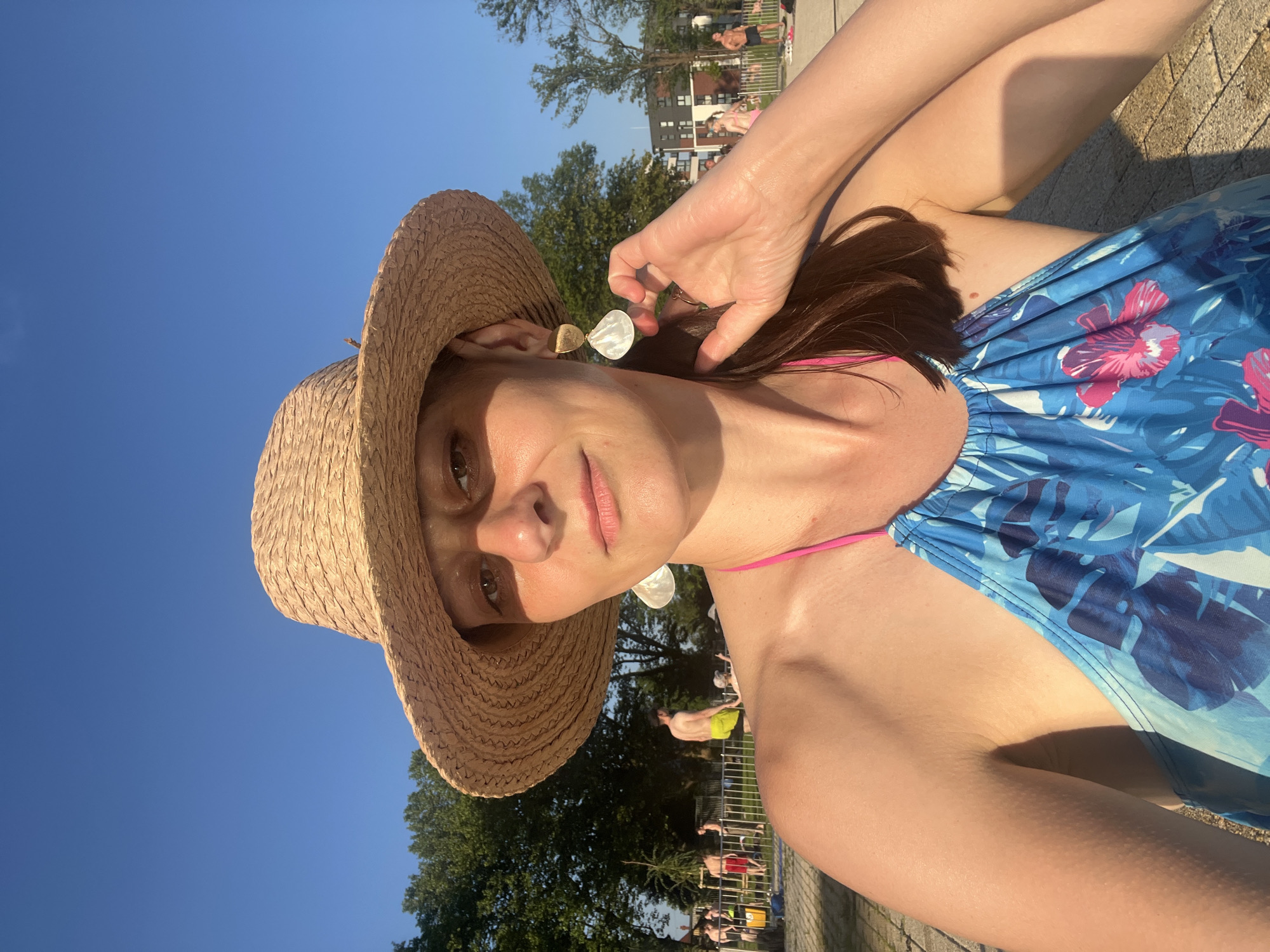 A woman wearing a straw hat and a blue swimsuit with a tropical print is posing for a selfie. She is outdoors with a clear blue sky and trees in the background. There are other people in the background, some of whom appear to be near a body of water, possibly a pool or beach. The lighting suggests it is daytime, likely afternoon, with the sun casting shadows.