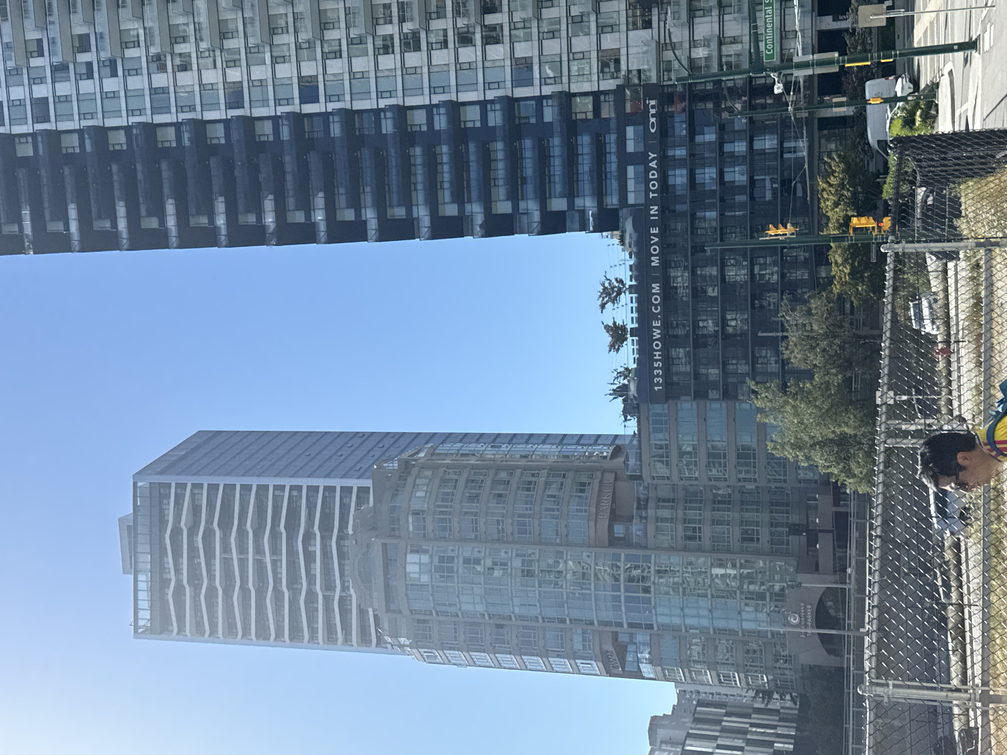 A sunny day in Vancouver, Canada, is captured in this image featuring the distinctive architecture of the city's downtown core. The scene is dominated by towering residential buildings, with a particularly striking glass-clad structure in the foreground exhibiting a curved facade and tiered balconies. To its right, a darker, more angular high-rise presents a stark contrast with its deep-set windows and narrow balconies. A billboard on the dark building advertises "335HOWE.COM | MOVE IN TODAY | Omni". In the middle ground, trees provide a touch of greenery against the urban backdrop. A chain-link fence runs along the bottom of the frame, separating the street from a pedestrian area where a person wearing sunglasses and a yellow and red shirt walks past, partially visible. Cars and a white van are also present in the background. Street signs indicate "Continental St".