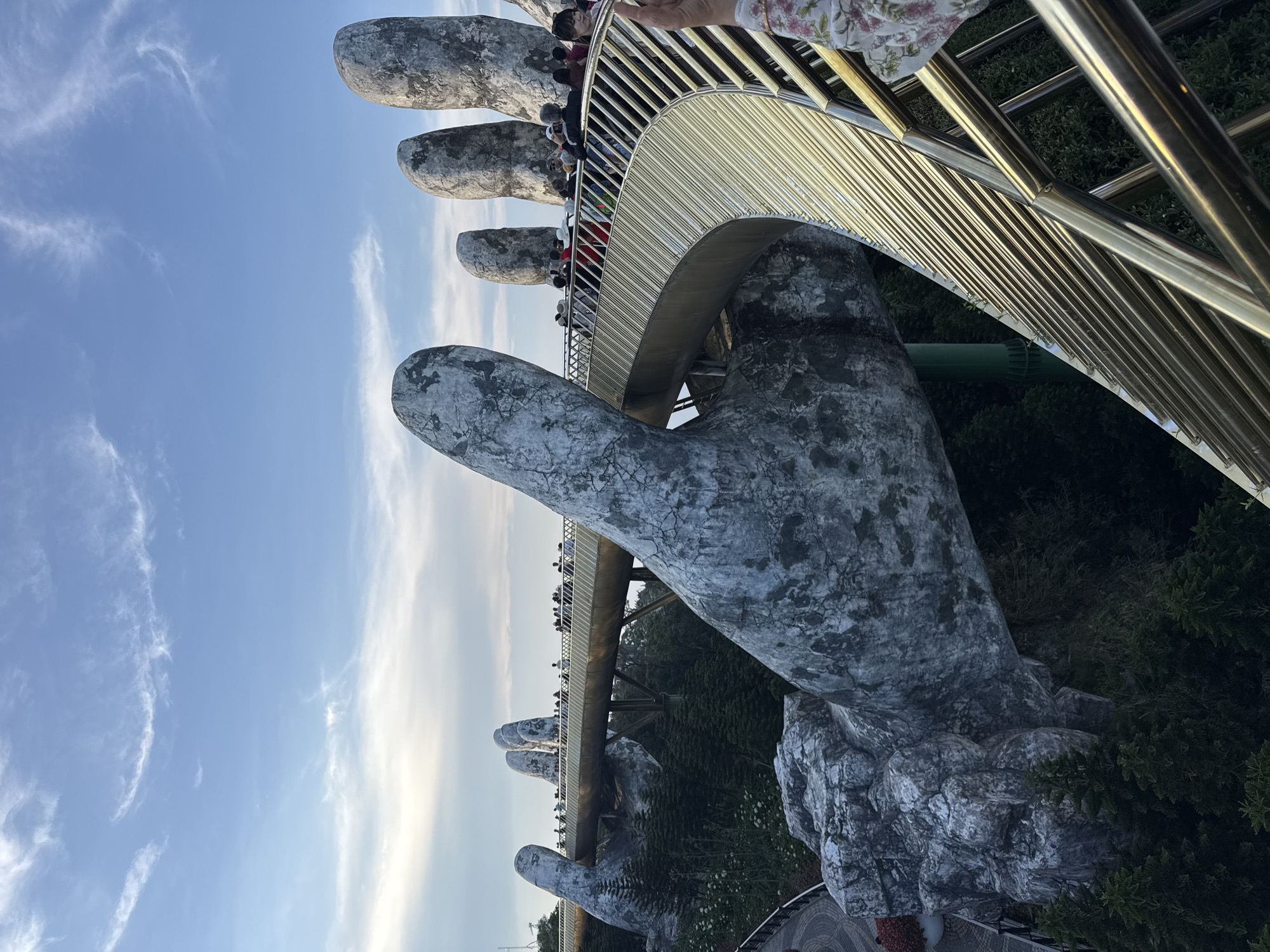 The image displays the Golden Bridge (Cầu Vàng) located in Bà Nà Hills, near Đà Nẵng, Vietnam. The central subject is a curved, golden-colored pedestrian bridge, held aloft by two enormous, weathered concrete hands designed to resemble ancient stone. The bridge features a golden-hued walking surface and robust gold-colored metal railings consisting of multiple horizontal bars, curving from the lower right foreground towards the upper left background.
Numerous individuals are visible walking along the bridge, appearing to be tourists. They are casually dressed and distributed along the structure, with a denser cluster on the right side of the frame. Their actions include walking and observing the surroundings. The giant thumb of one hand structure dominates the foreground, reaching up to support the bridge. Further along, the fingers of the second hand are clearly visible, cradling the bridge deck. In the background, additional rounded, stone-like structures, resembling more fingertips, are silhouetted against the sky.
Below the bridge, a dense canopy of dark green coniferous trees and other foliage covers a steep hillside, indicating a mountainous environment. The sky above is a gradient of clear blue transitioning to a lighter, hazy blue near the horizon, with sparse, wispy cirrus clouds. The lighting suggests late afternoon, with sunlight reflecting off the golden railings and illuminating the scene. A partially visible arm and patterned sleeve in the extreme upper right foreground indicate the photographer's close proximity to the bridge railing.