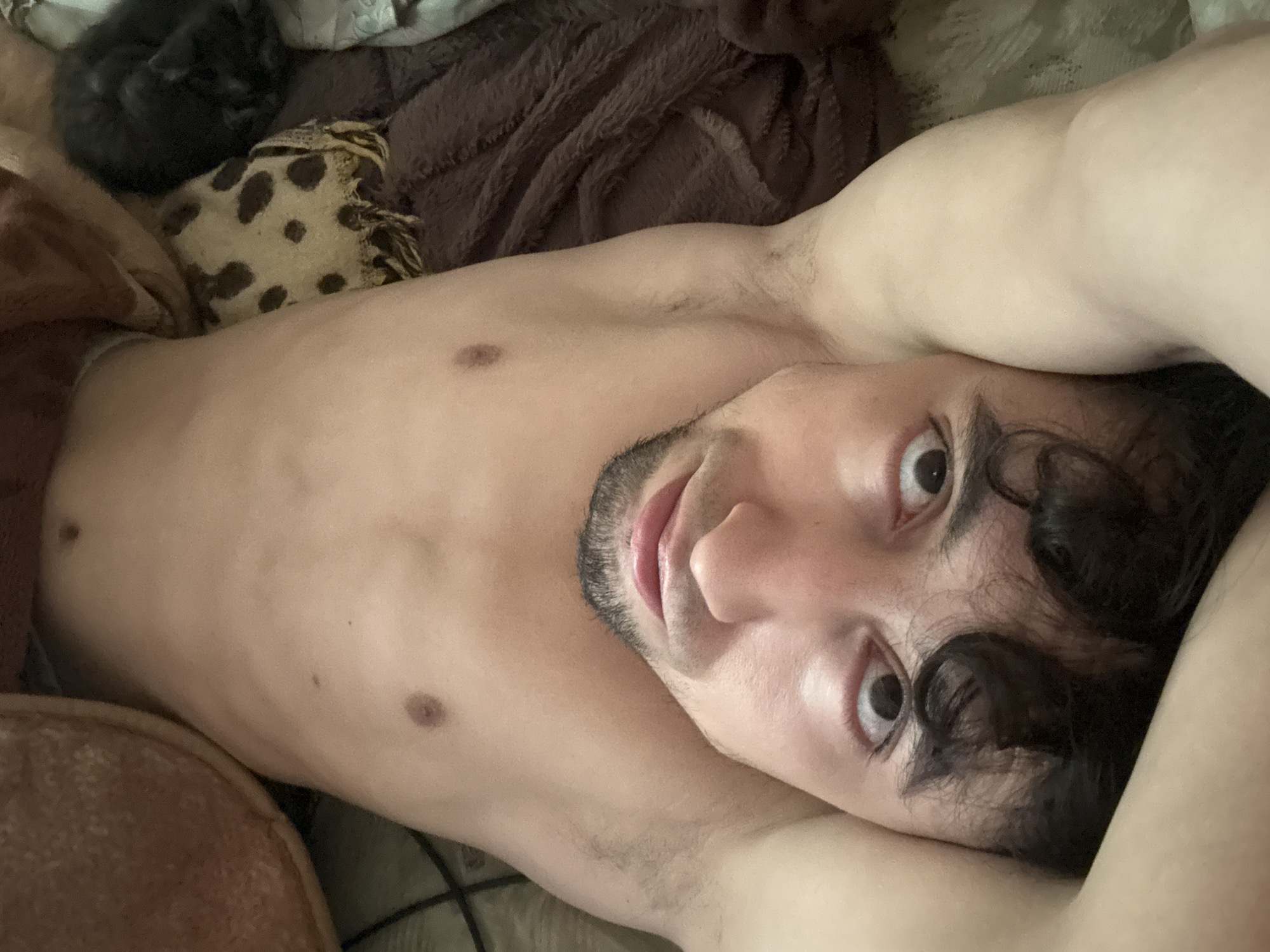 An overhead, upside-down, close-up selfie shows a bare-chested individual lying on their back. The person's head is at the bottom of the frame, with their eyes wide open and looking at the camera. They have dark, curly hair, a mustache, and a light beard. Their torso is visible, with no shirts or pants. The individual is on a bed with brown blankets and a patterned pillow. A small black cat is visible on the right side of the frame, curled up on a brown blanket. The background appears to be a bedroom.