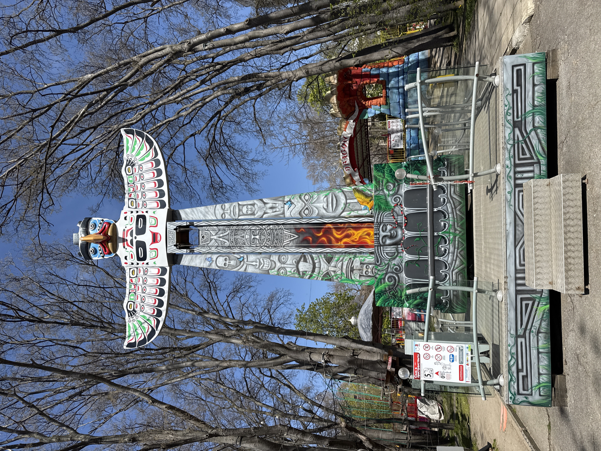 The image captures an amusement park scene under a clear, bright blue sky, framed by numerous bare-branched trees, suggesting late winter or early spring.
The central subject is a tall, colorful amusement park ride resembling a totem pole. At its apex is a large, stylized bird figure with outstretched wings, predominantly painted in white, black, red, and green, with blue, red, and yellow accents on its face. The ride's central column features intricate gray and white tribal patterns, including stylized faces and geometric designs, with a prominent section displaying orange and red flame-like imagery.
To the right of the main ride, there's an entrance area equipped with metal stairs and railings. The base of the ride and the surrounding structure are adorned with a vibrant mix of gray, black, and green artwork, featuring tribal patterns and stylized jungle foliage. A white sign near the entrance provides safety information and regulations, utilizing pictograms to indicate restrictions for pregnant women, small children (age 0-3 indicated with a crossed-out symbol), individuals with certain health conditions, and dogs. A red downward-pointing triangle with the number "5" is also visible. Text on the sign, in Bulgarian, includes "Внимание!" (Caution!).
In the background, parts of other amusement park structures are visible, including one with a red and blue block design and the word "DRAGON" painted in red and white. The ground beneath the structures is paved asphalt. No people are visible, giving the impression of a quiet or off-season amusement park in Varna, Bulgaria.