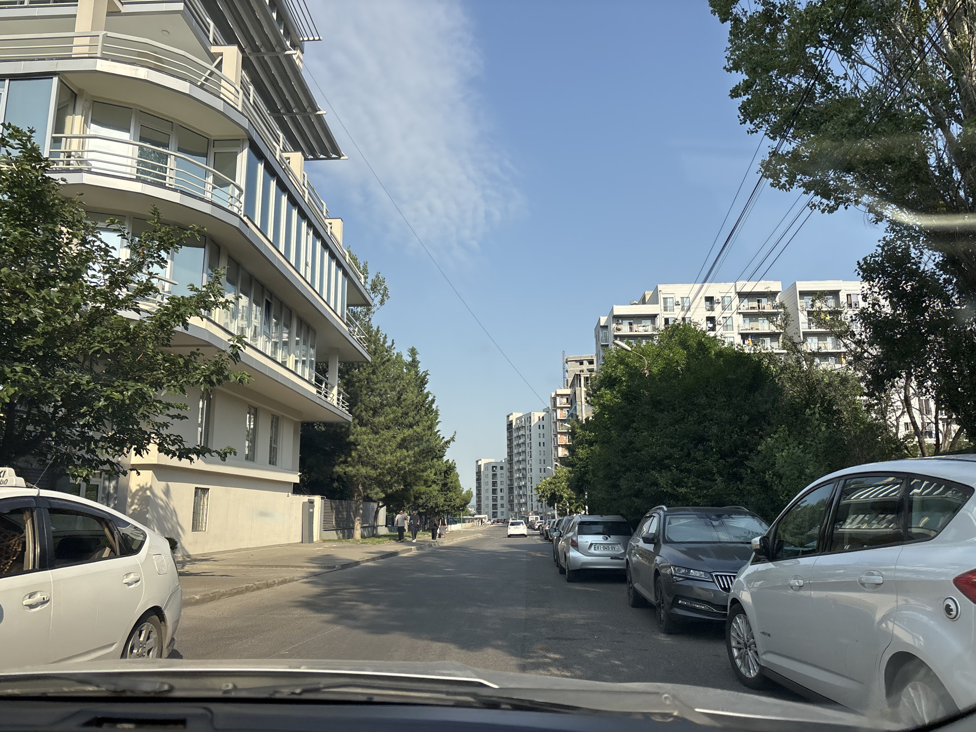 The image depicts a street scene in Tbilisi, Georgia. On the left side of the road, a modern, multi-story building with curved balconies and large windows dominates the foreground. Several trees line the sidewalk in front of this building. A white taxi is parked along the sidewalk. The road is asphalt and appears to be a two-way street with cars parked on the right side. Further down the street, several other modern apartment buildings are visible, some under construction. The sky is blue with some scattered white clouds. Several people are walking on the sidewalk to the left, and cars are parked along the right side of the street. Overhead, electrical wires are strung across the scene. The perspective suggests the photo was taken from inside a vehicle.