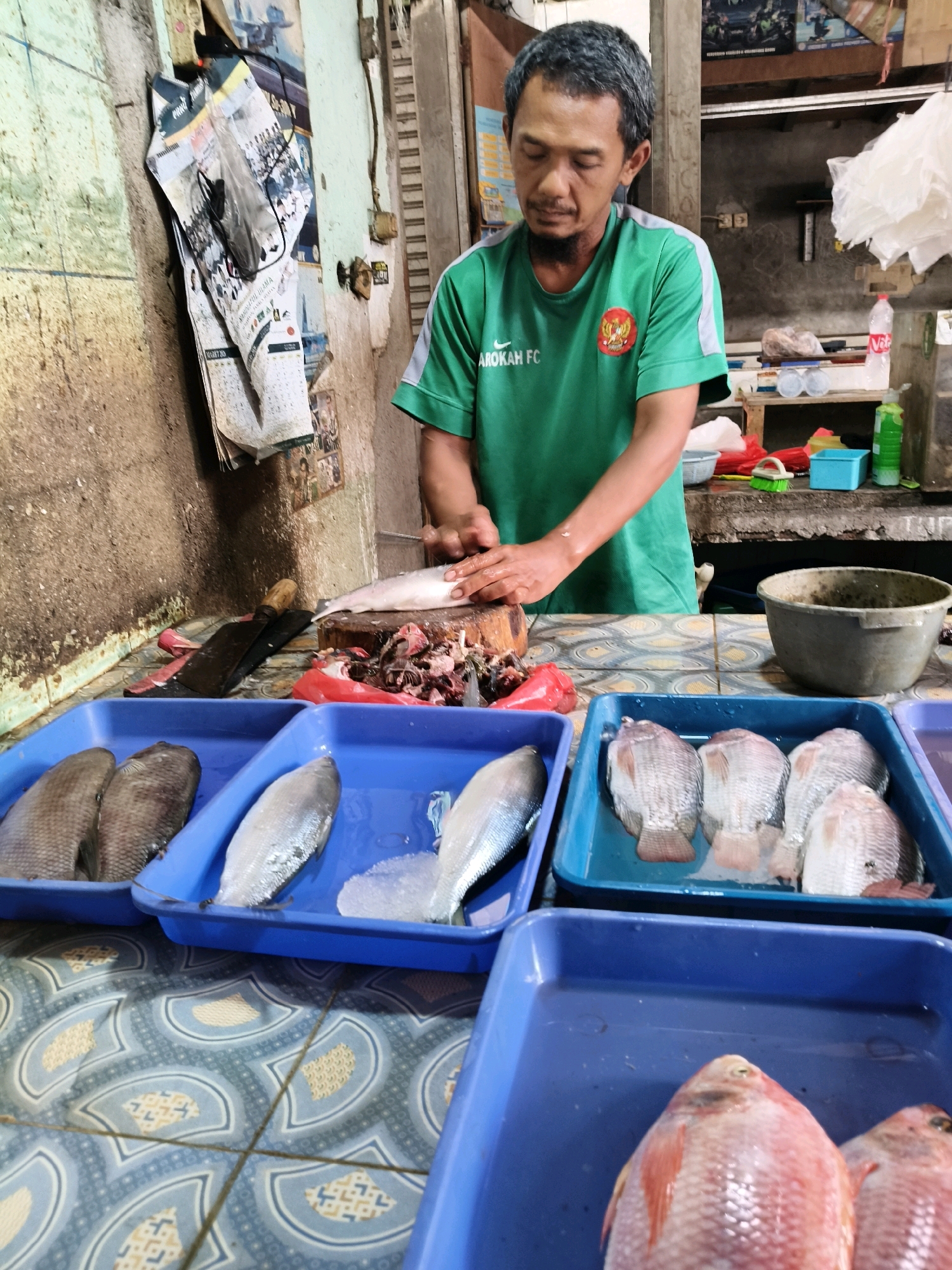 A man is seen standing at a fish market stall in Setiamekar, Indonesia, preparing fish. He is wearing a green t-shirt with a logo and gray accents. He is in the process of cutting a fish on a wooden chopping board with a large cleaver. To his left, several fish are laid out in blue plastic trays. In front of him, there are more blue trays containing various types of fish, including tilapia. Behind him, there is a wall with posters and calendars, and shelves with containers and plastic bags. The stall appears to be outdoors or in an open-air market. The overall scene depicts a typical day at a local fish market.