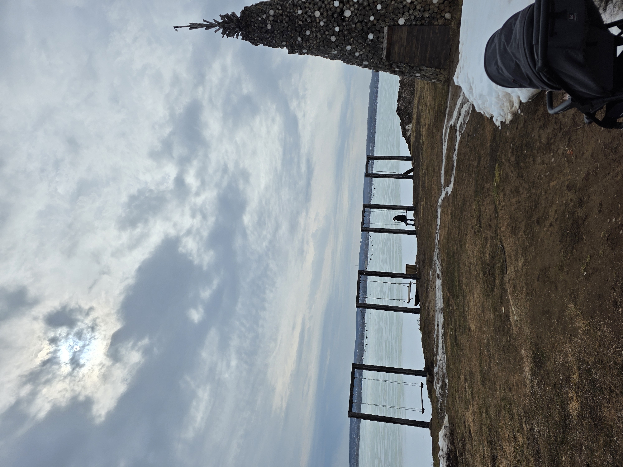The image captures a scene at the Votkinsk Reservoir in Votkinsk, Russia. The day is overcast, with a cloudy sky and the sun partially obscured. The reservoir extends into the horizon. Along the shore, there are several wooden swing sets. A person is sitting on one of the swings. To the left, a decorative structure resembling a tree built from logs and white spheres is visible. Adjacent to it, on the right, there is a patch of snow and part of a baby stroller. The ground is covered with grass and dirt, showing signs of melting snow.