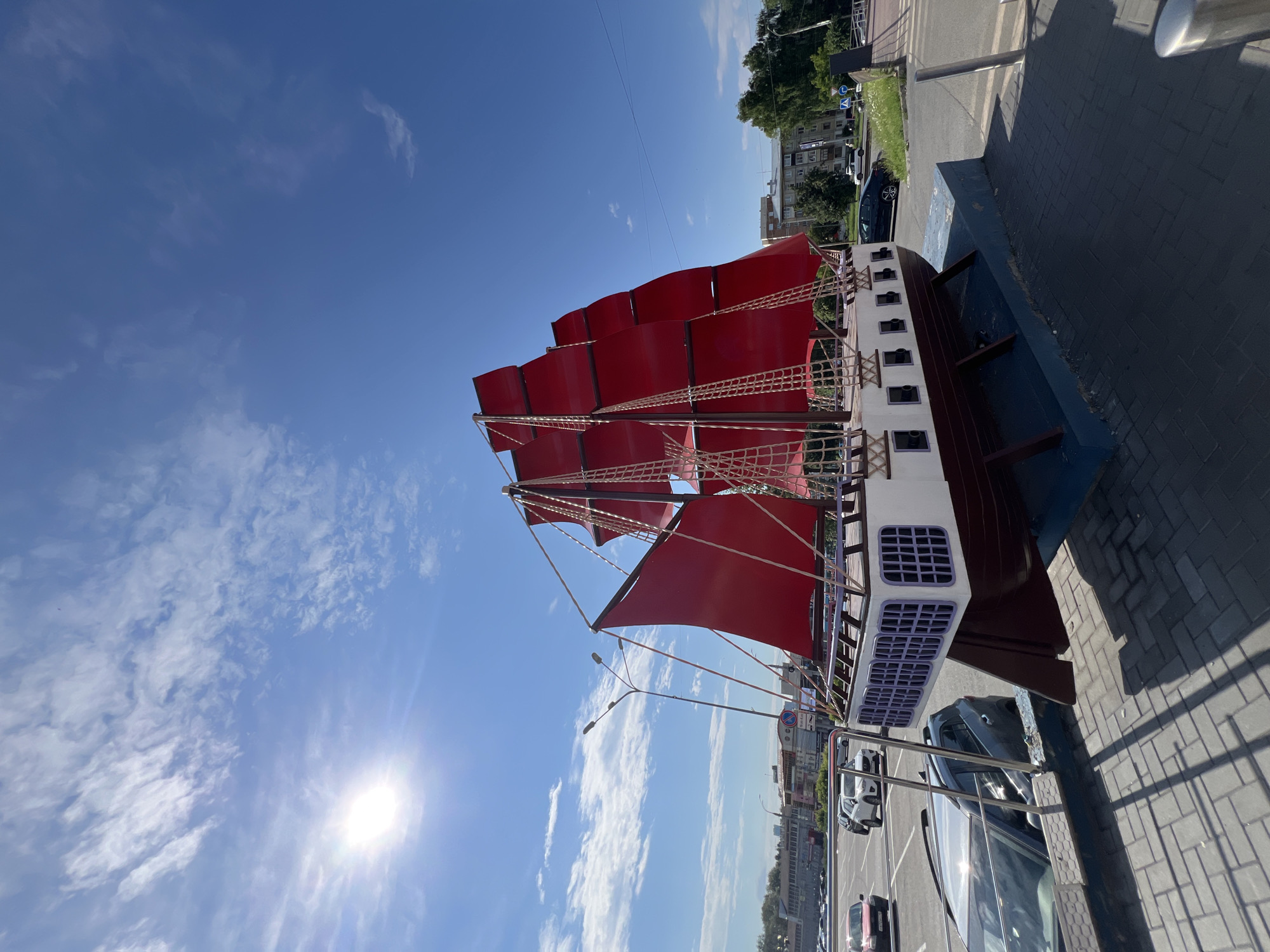 The image shows a large, red-sailed ship model displayed outdoors on a sunny day in Izhevsk, Russia. The ship is the central focus, positioned on a blue pedestal with wooden supports. It has white and maroon detailing along its hull, and a deck with railings and what appear to be windows. Red fabric sails are stretched taut on masts and rigging. In the background, a street with cars is visible, including a silver SUV and a red car parked in a lot. Beyond the street, buildings and trees form the urban landscape. The sky is a clear blue with scattered white clouds, and the sun shines brightly. The ground in the foreground is paved with gray bricks, casting distinct shadows. The overall scene suggests a public space, possibly a park or a prominent roadside attraction in the city of Izhevsk.