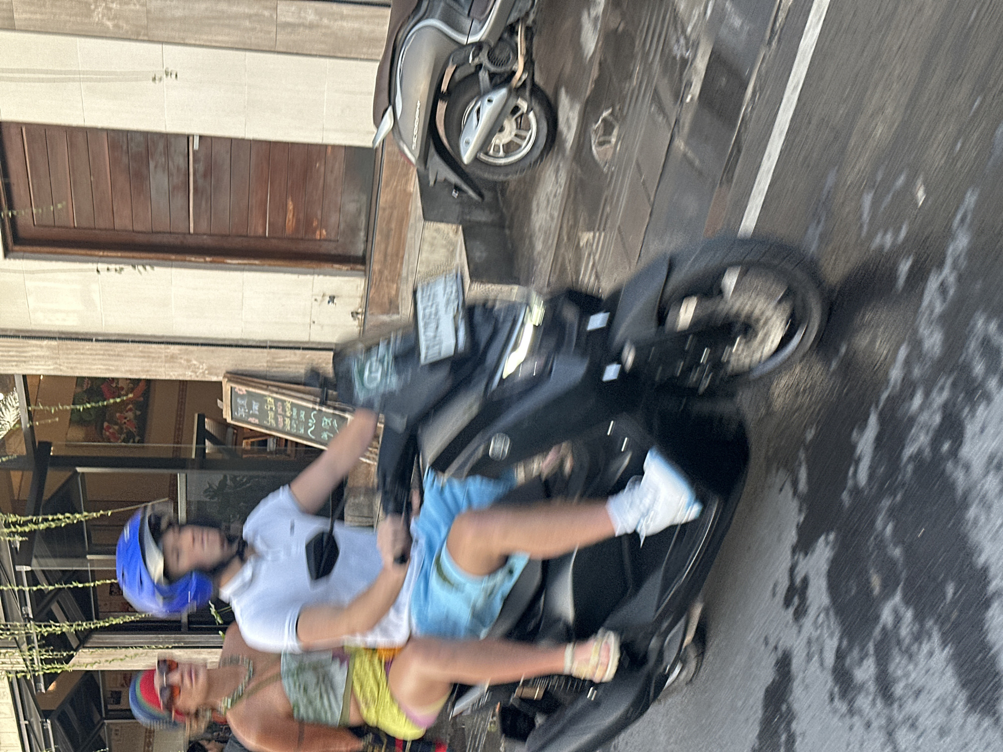 In North Kuta, Indonesia, two individuals are riding a dark-colored scooter on a wet asphalt street. The person in the front, wearing a blue helmet and a white polo shirt with blue shorts, is driving. The passenger behind them, with a colorful headband and sunglasses, wears a strapless green and yellow patterned top and yellow shorts. The street has a white line and reflects the overhead lights. In the background, a building with a wooden door and light-colored tiled walls is visible, along with hanging plants and a sign with text. Another scooter is parked to the right. The image is slightly blurred due to motion.