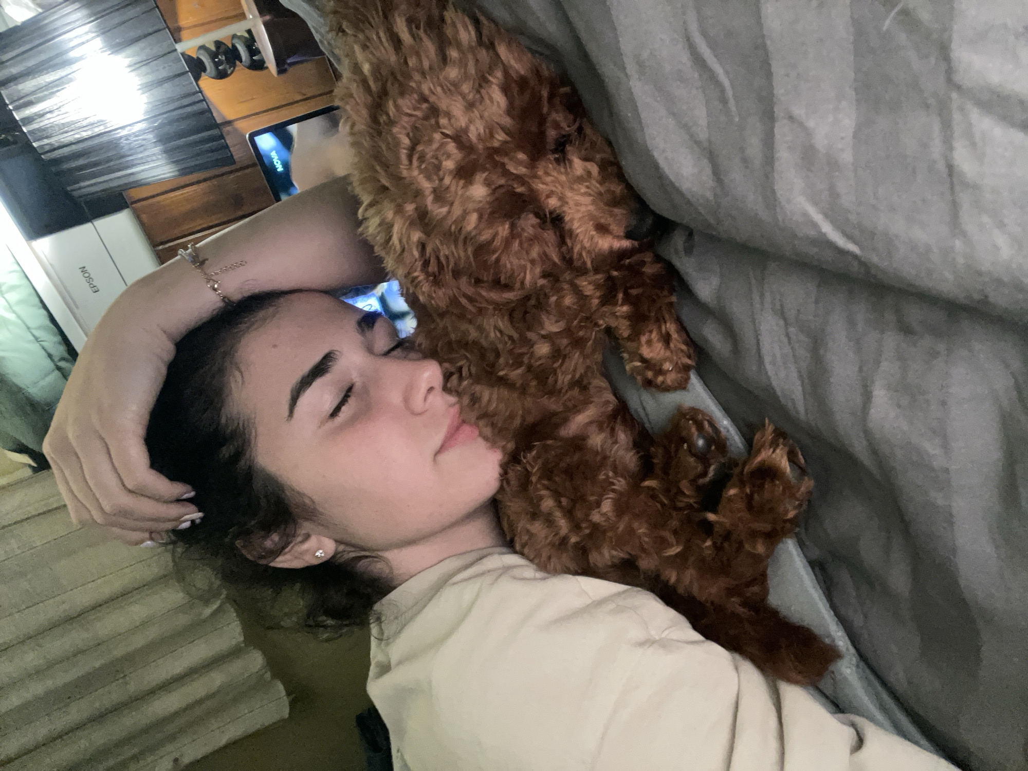 A young woman with dark curly hair, wearing a light-colored shirt, lies on a gray bed with her eyes closed. Her left hand rests on her head, and a small earring is visible in her left ear. A brown, curly-haired dog is lying next to her, partially on top of her body and under the gray bed sheets. An Epson printer and a wooden dresser with items on top are visible in the background. There is no visible evidence suggesting the location of Sofia, Bulgaria.