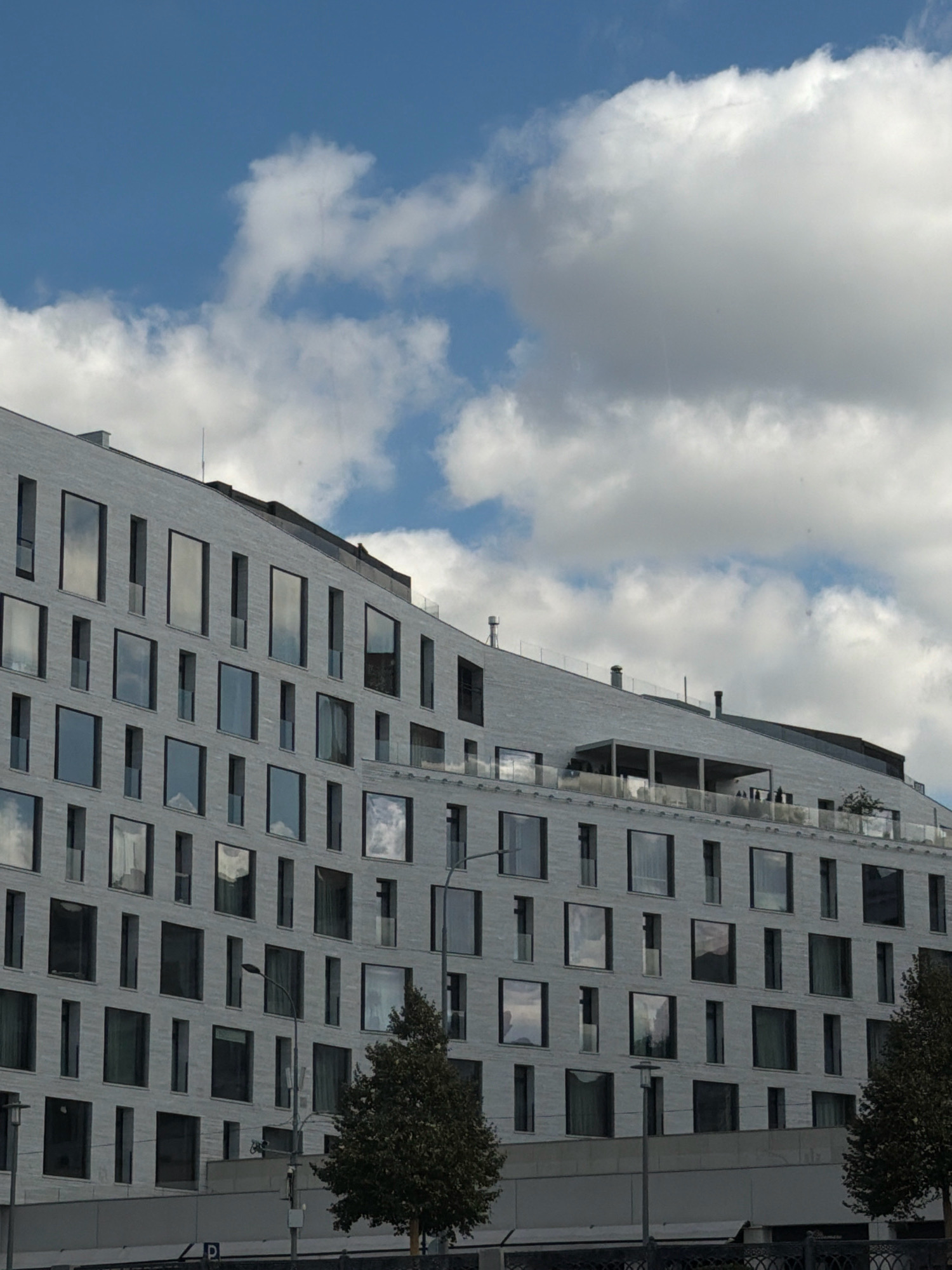 The image depicts the facade of a modern, multi-story building, possibly a hotel or residential complex, in Moscow, Russia. The building features a light gray, textured exterior and a series of large, dark-framed windows arranged in a geometric pattern. Some windows reflect the cloudy sky, which is a prominent element in the upper portion of the image, featuring white cumulus clouds against a blue sky. The building's roofline is angled, and it appears to have multiple levels with balconies or terraces. In the foreground, partially visible, are streetlights and trees, suggesting an urban environment. The overall impression is one of contemporary architecture set against a dynamic sky.