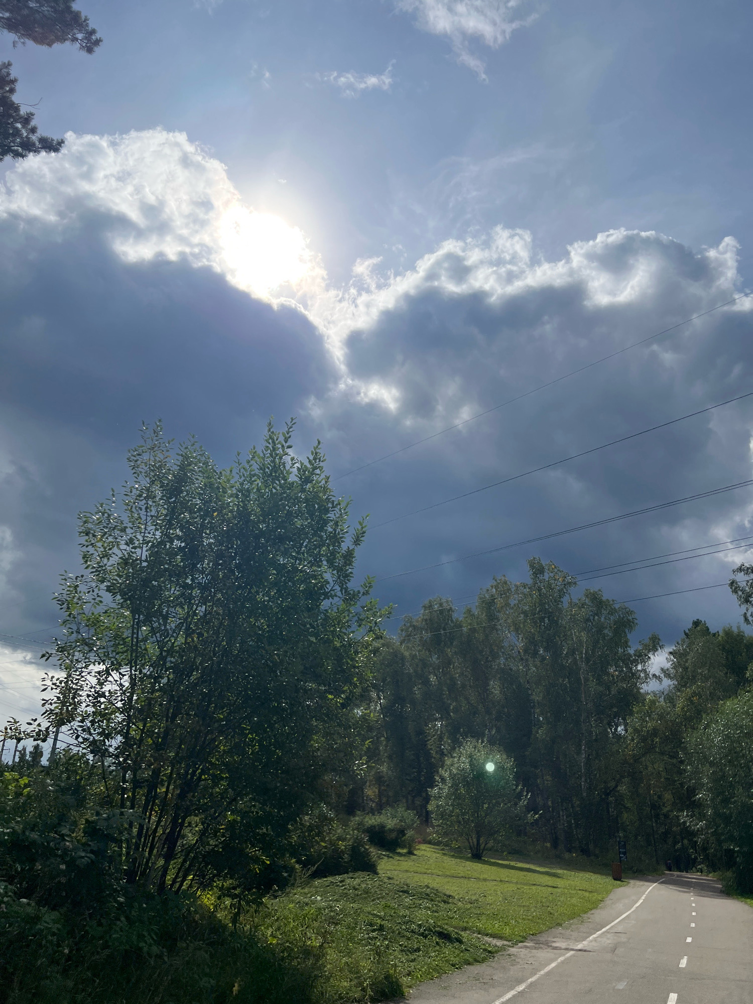 The image depicts a scenic view of a paved pathway winding through a park-like setting, likely within the city of Krasnoyarsk, Russia, given the presence of birch trees and the general landscape typical of Siberia. The pathway, marked with white dashed lines, curves to the right and recedes into a dense forest. To the left of the path, a grassy slope is dotted with small trees and bushes. Overhead, a dramatic sky unfolds with a mix of dark, heavy clouds and patches of bright sunlight peeking through. A few power lines are visible stretching across the upper portion of the frame. The overall impression is one of natural beauty with hints of infrastructure. No people are visible in the frame.