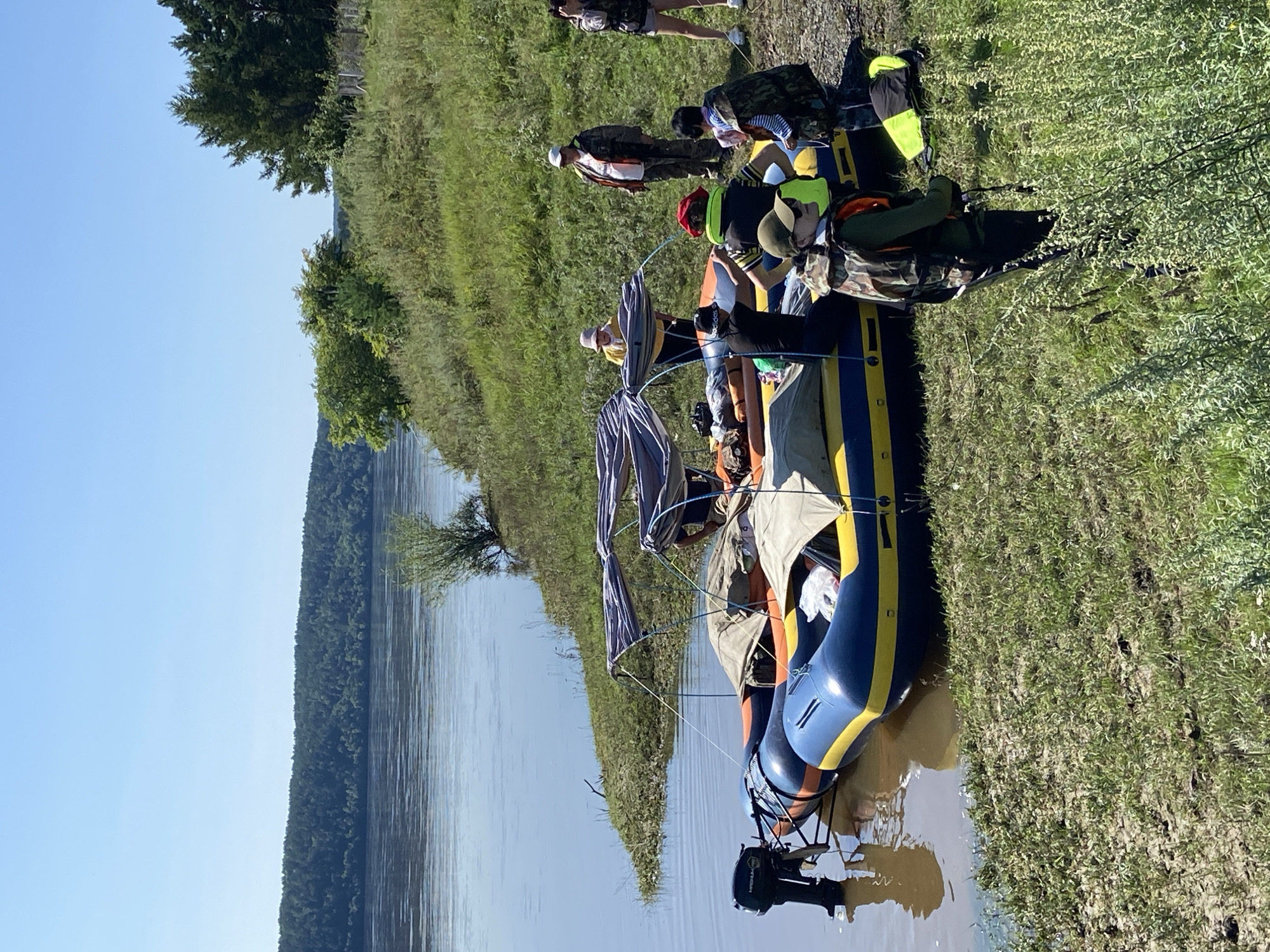 A group of people are gathered near an inflatable raft on the shore of a wide river. The raft is partially covered with tarps and has a small outboard motor attached to the back. The individuals are dressed in casual clothing, with several wearing life vests. The environment is a grassy embankment with trees and a forest visible in the background across the river. The sky is clear and blue, indicating a sunny day.