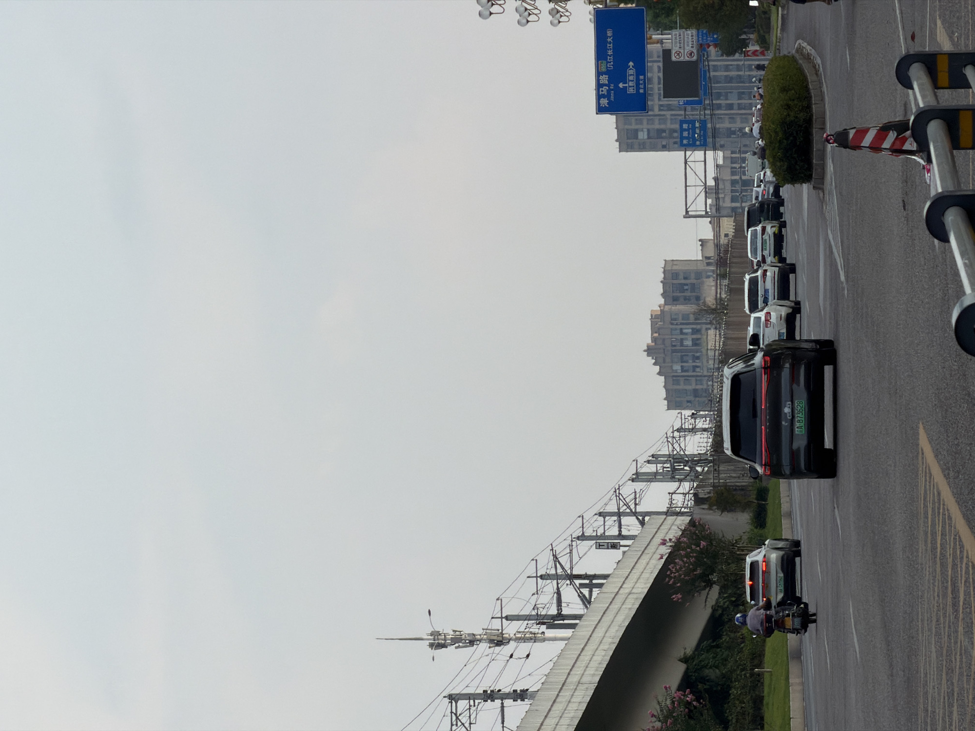 The image depicts a street scene in Jiangjin, China, under an overcast sky. A multi-lane road is occupied by several vehicles, including a motorcycle with a rider, a dark SUV, a lighter SUV, and multiple sedans. The road appears to be an elevated highway or overpass, indicated by the presence of concrete barriers and overhead power lines supported by metal poles. In the background, modern buildings are visible, suggesting an urban environment. A blue traffic sign with Chinese characters and arrows points towards "津马路" (Jinma Road) and "长江大桥" (Changjiang Bridge). The overall atmosphere is that of a typical day in a busy Chinese city.