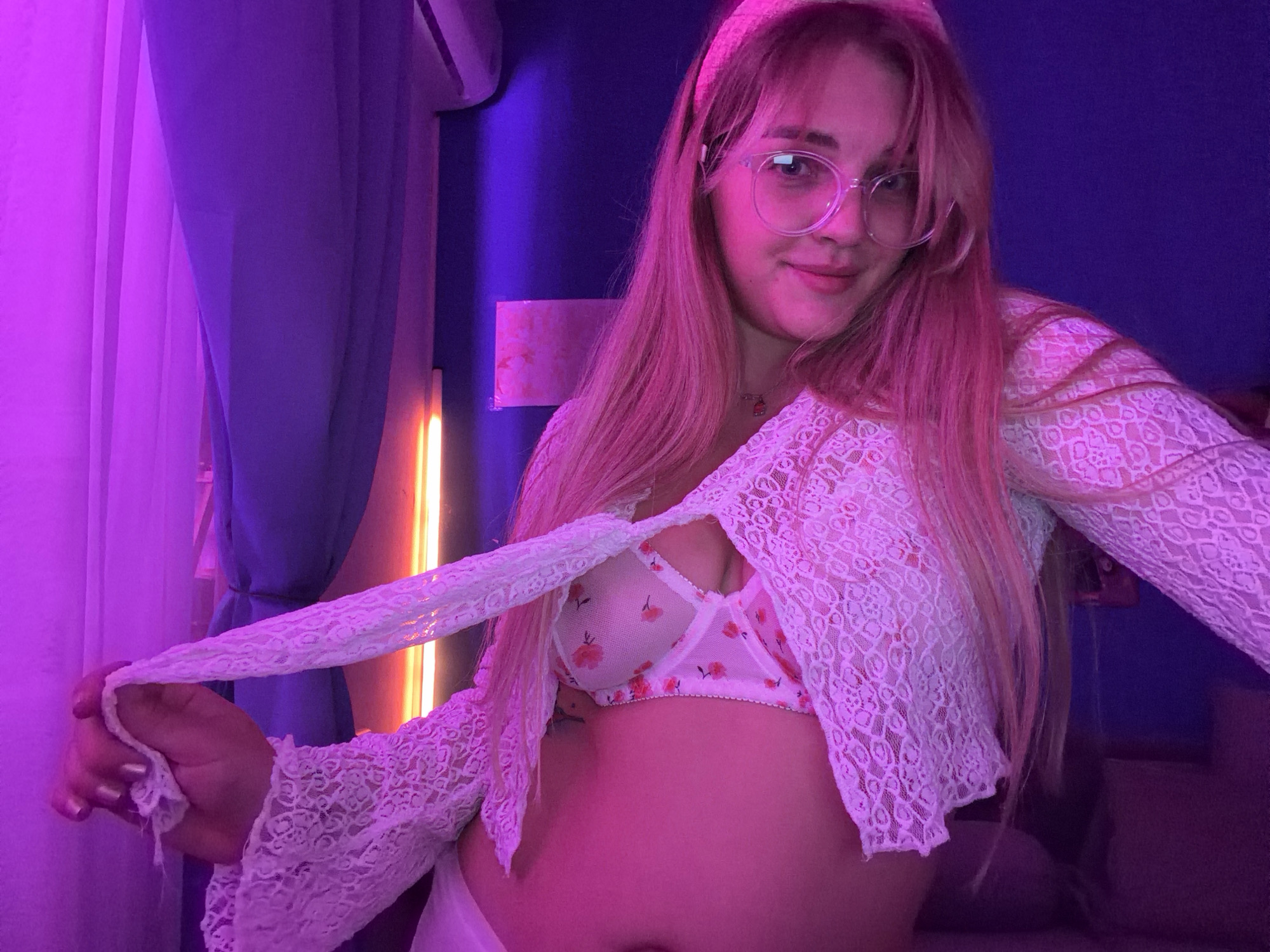 A young woman with pink-tinted blonde hair and clear-framed glasses is the primary subject, facing the camera with a slight smile. She wears a white lace top, partially open to reveal a white bra adorned with a floral pattern. Her skin appears to have a pinkish hue, likely due to the lighting in the room. She holds a piece of the lace fabric in her left hand.
The background features a purple curtain on the left and a blue wall behind her. A vertical light fixture is visible near the curtain. The setting appears to be an indoor room, possibly a bedroom, with other furniture or items partially visible in the periphery. The location is specified as Большая Шильна, Russia, but the image itself provides no independent verification of this geographical claim.
