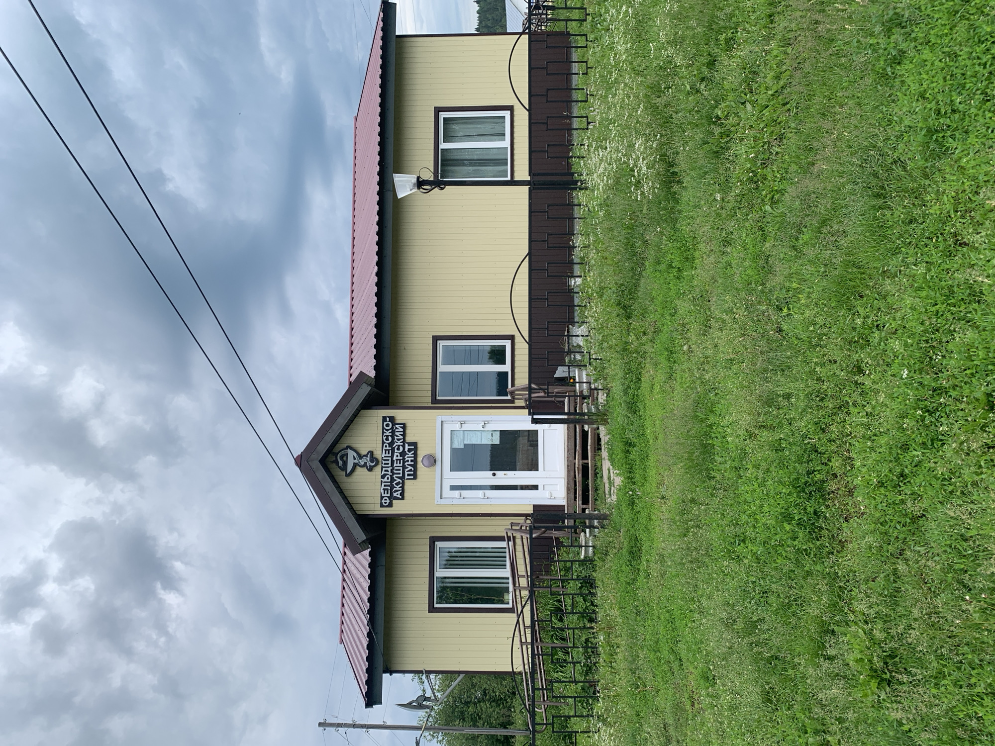 The image shows a small, light yellow building with a maroon roof, labeled "ФЕЛЬДШЕРСКО-АКУШЕРСКИЙ ПУНКТ" which translates to "Feldshersko-Obstetric Point" in Russian. The building has two windows on the left side and two on the right side of the entrance, which consists of a double white door. A ramp for accessibility is visible to the left of the entrance. A black metal fence with arched patterns separates the building from a grassy area in the foreground, which is covered in green grass and some small white flowers. A black lamppost is positioned in front of the right side of the building. The sky above is cloudy and overcast. Power lines are visible stretching across the upper portion of the image. The overall scene suggests a rural or semi-rural setting, possibly a medical facility serving a small community in Barsai, Russia.