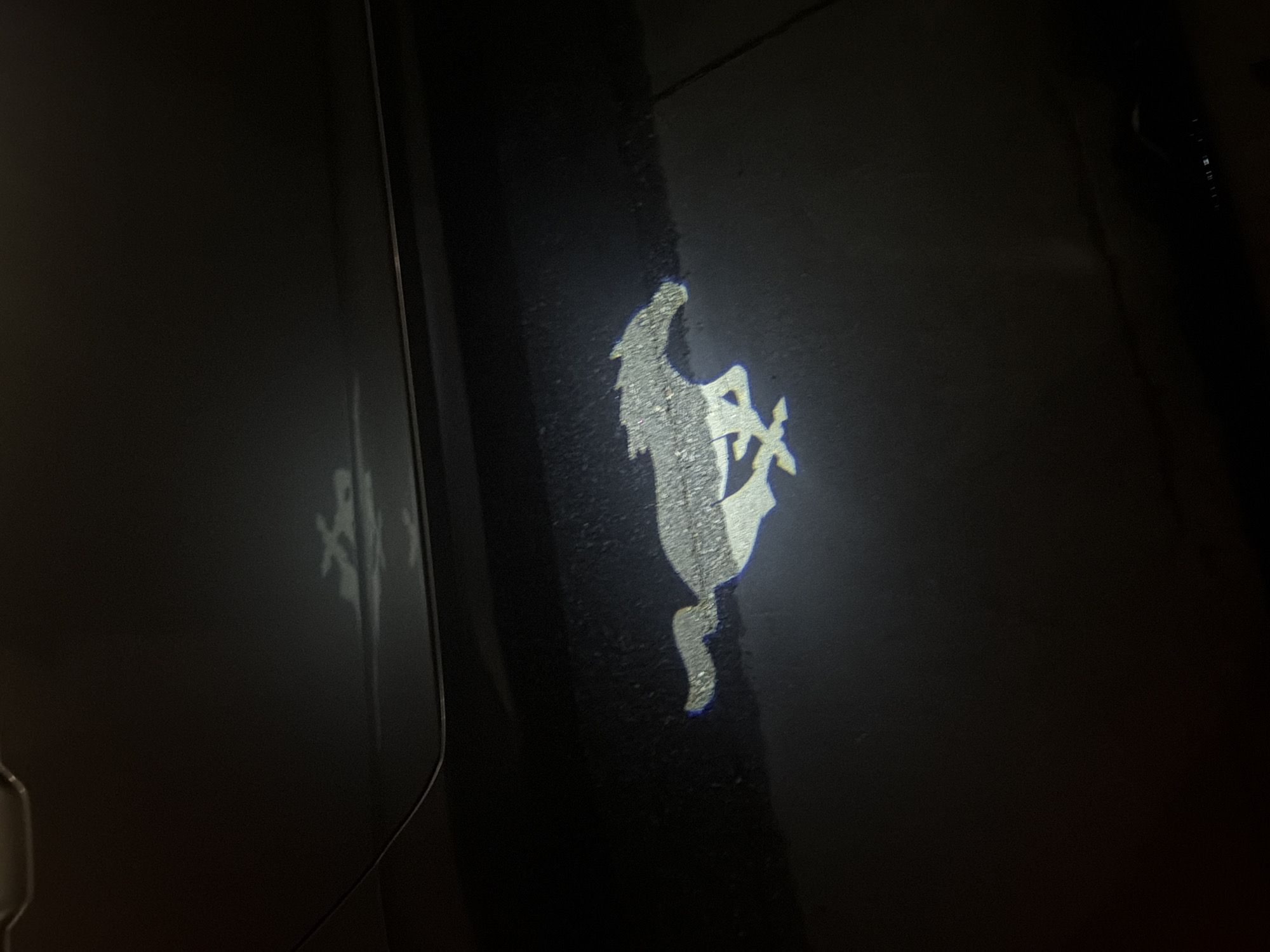 A projection of a prancing horse emblem is cast upon a dark surface, likely a vehicle door. The emblem is depicted in a bright, possibly white, light, contrasting against the dark background. The surface appears to be wet or glossy, reflecting the projected image to a fainter degree on a neighboring area. The scene is dimly lit, with minimal ambient light contributing to the overall darkness. No people or identifiable objects are visible other than the surface onto which the projection is cast. The location in Palm Desert, United States of America cannot be discerned from the image.