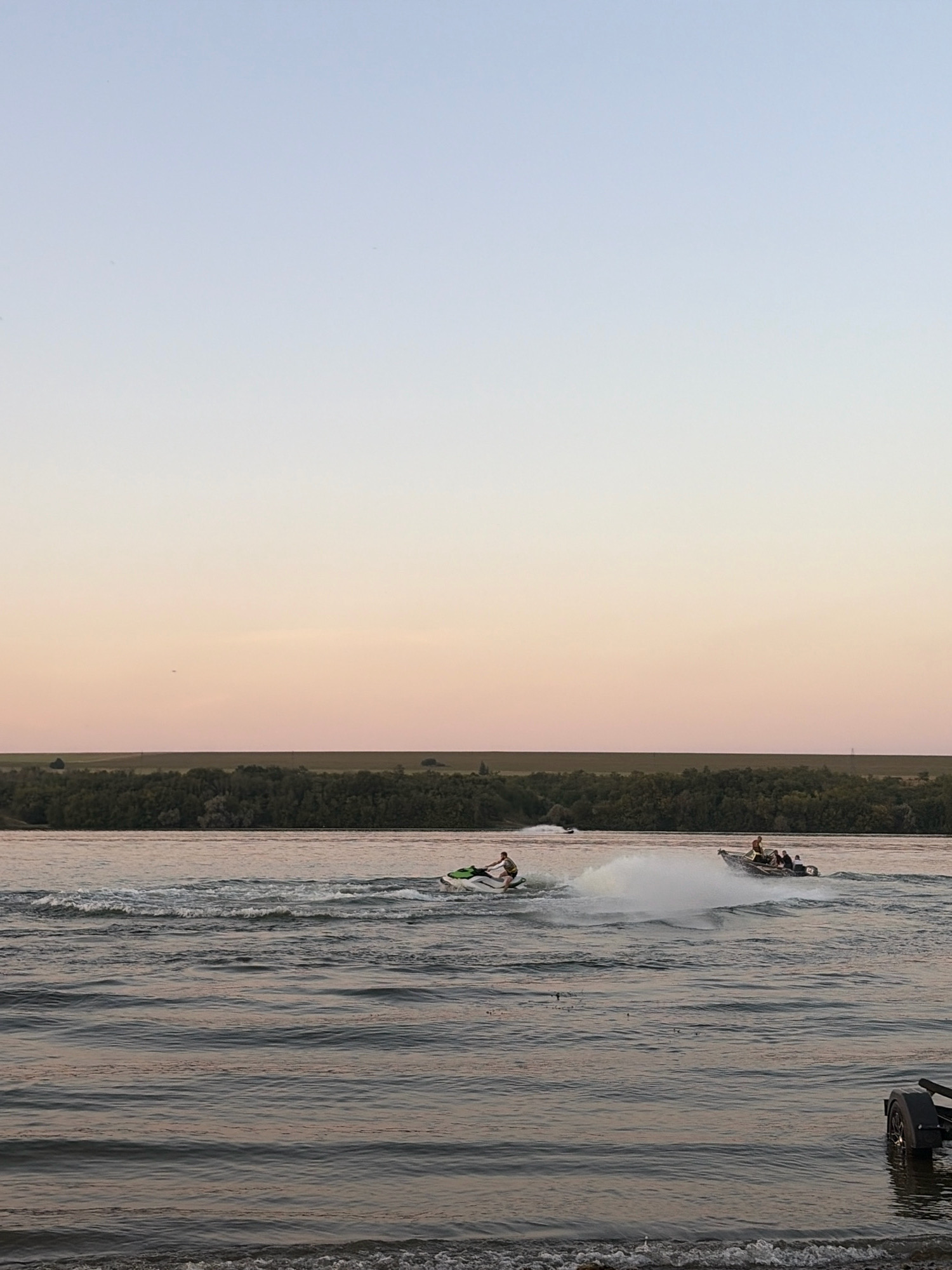 In Oxentea, Moldova, two personal watercraft and one motorboat are active on the Nistru River. On the left, a person rides a jet ski, leaving a white wake. To the right, a motorboat with three people aboard is generating a large spray of water as it moves at speed. A second jet ski is visible in the distance behind the motorboat. The river reflects the soft hues of the twilight sky, transitioning from pale blue to a soft pink. The far bank of the river is lined with a dense band of green trees, beyond which a flat landscape stretches towards the horizon. The foreground shows the edge of the river with small waves lapping at a gravelly shore, and a portion of a trailer is visible on the right.