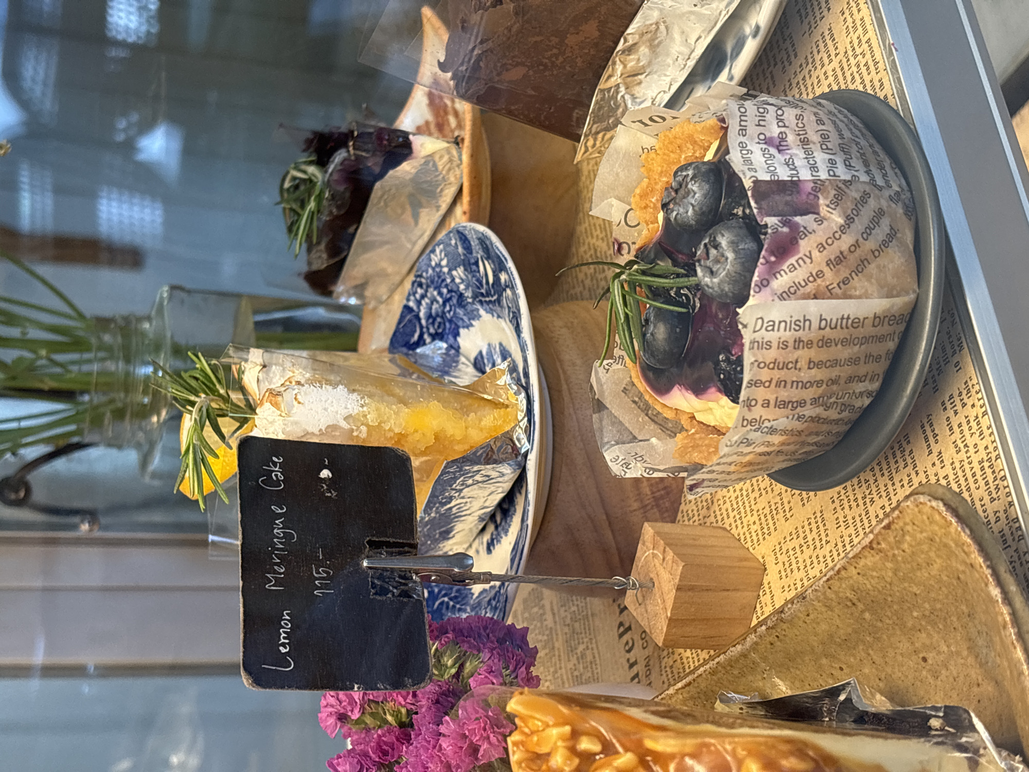 This image shows a display case of baked goods, likely in a cafe or bakery. A sign labeled "Lemon Meringue Cake - 115.-" identifies one offering. The cakes and tarts are arranged on a wooden display platform. Cakes include blueberry and possibly chocolate. The food items are decorated with garnishes such as flowers and herbs. The environment suggests an establishment located in Choeng Thale, Thailand based on the language displayed.