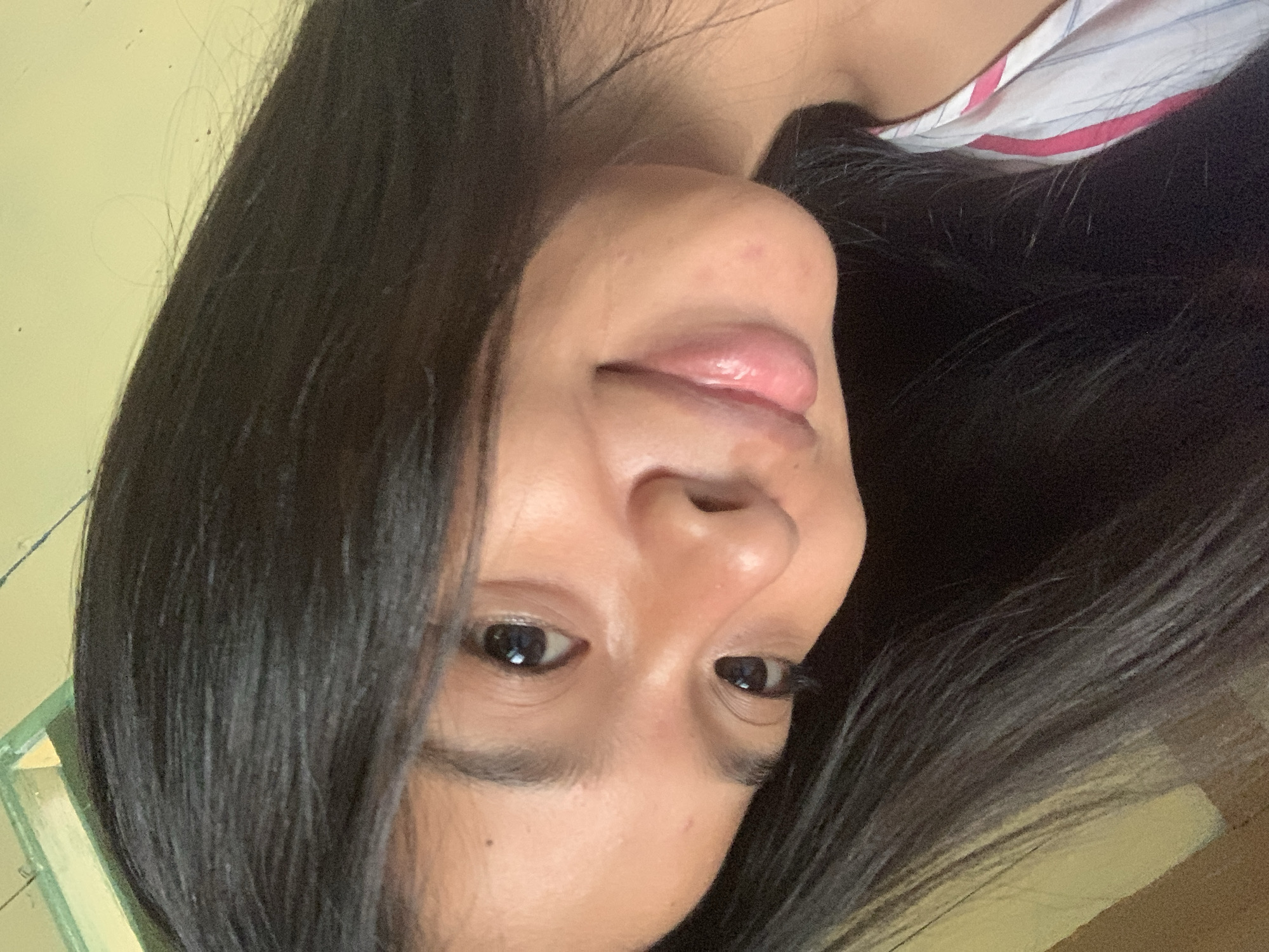 A close-up, slightly tilted selfie shows a young woman with long, dark hair. Her head is tilted to the left, and she is looking towards the camera. Her eyes are dark, and she has a gentle smile on her face, with her lips slightly parted. Her skin is smooth and has a warm, tan complexion. She is wearing a striped shirt with pink and white colors. The background appears to be a wall with a yellow hue and a green painted frame at the top left corner. The image suggests an indoor setting, possibly a home environment. There is no visible indication of the specific location in Sungai Meriam, Indonesia.