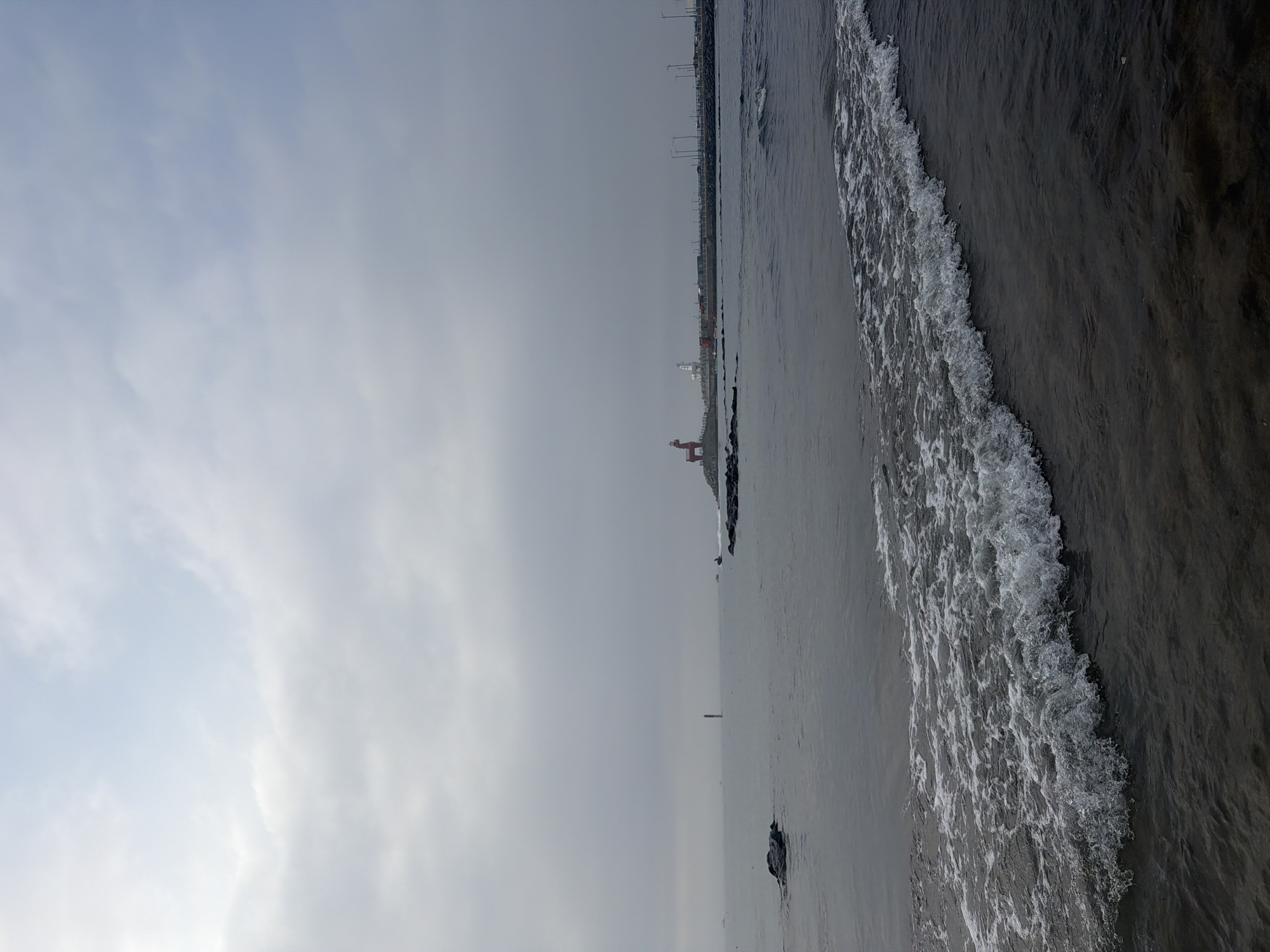 The image shows a view of the ocean near a harbor on Jeju Island, South Korea. In the distance, a red reindeer-shaped structure and a lighthouse are visible on a pier. The water in the foreground is dark and choppy, with a wave breaking on the shore. The sky is overcast with a pale blue hue. The scene is devoid of any people.