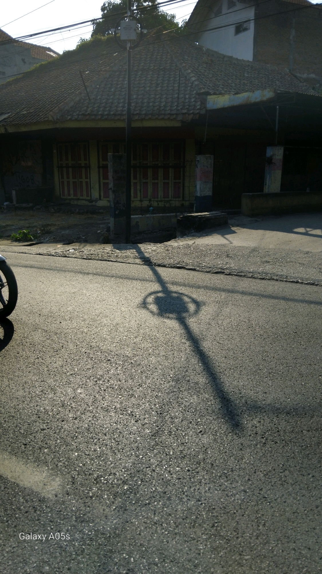 The image shows a street scene in Sumedang, Indonesia, during daylight hours. A street lamp casts a distinct shadow on the asphalt road, with a circular fixture visible at the top of the shadow. To the left, a portion of a motorcycle wheel is visible. In the background, a building with a tiled roof and closed windows stands adjacent to the street. The area appears to be a mixed-use urban environment.