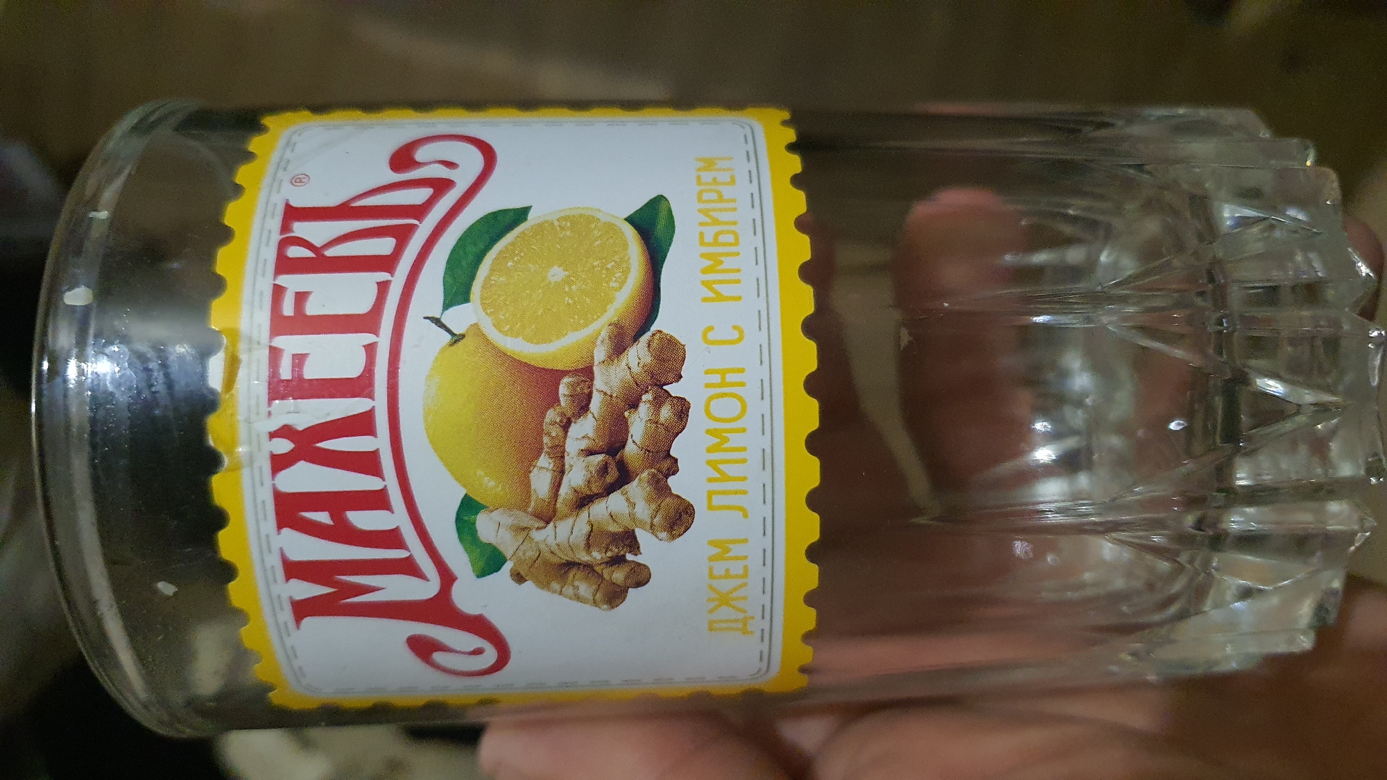 A close-up, eye-level shot shows a glass jar with a label that reads "МАХЕЕВЪ" in red lettering. Below the brand name are illustrations of lemons and ginger roots, with the text "ДЖЕМ ЛИМОН С ИМБИРЕМ" in yellow lettering. The jar is partially filled with a clear liquid. The background is blurred and appears to be a dimly lit indoor setting. The image does not provide any information that would suggest a location in Karankul, Uzbekistan, or any other specific city.
