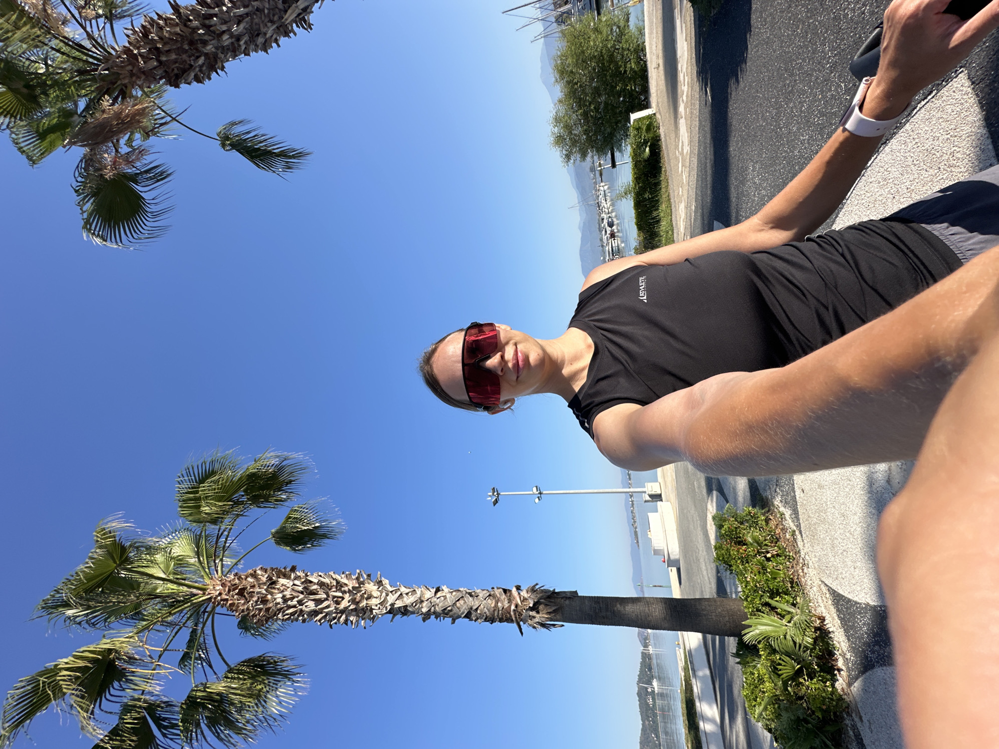 This selfie image, taken outdoors on a bright, sunny day, is notably rotated clockwise, giving the impression of an upward-looking shot with elements appearing horizontal or tilted. The dominant feature is a clear, vibrant blue sky filling a large portion of the frame.
The subject is a woman, partially seen on the right side of the image. She has fair skin, with her dark hair pulled back, and wears striking red mirrored sunglasses that obscure her eyes. Her expression is neutral. She is dressed in a black sleeveless athletic top, upon which the white, vertically oriented text "ATHLETE" is visible on her left chest. A white smartwatch is worn on her left wrist. Her arm extends into the foreground, suggesting she is holding the camera for the selfie.
The scene is a picturesque coastal environment, identified as Fethiye, Turkey. In the background, a marina is visible with numerous white boats and yachts docked on clear blue water. Distant mountains rise from the horizon across the bay. A paved road or promenade runs alongside the water, lined with green bushes and at least two palm trees. One large palm tree with a thick, textured trunk and green fronds extends from the lower right foreground, while another is partially visible on the far left. The strong, direct sunlight suggests it is daytime, likely mid-morning or afternoon, given the intensity of the light and the clear shadows. The weather is sunny and cloudless, contributing to the bright, summery atmosphere.