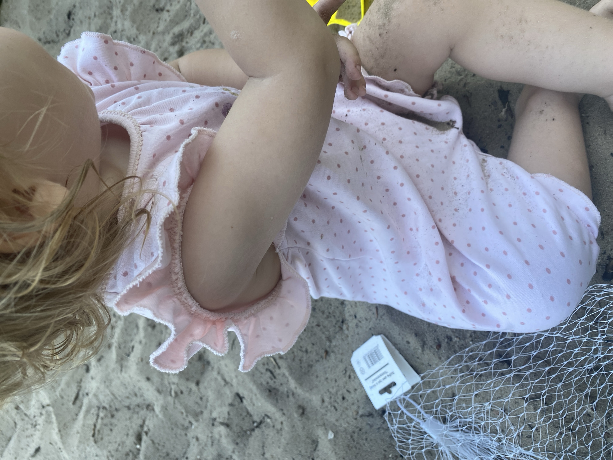 The image shows a close-up of a young child's arm and torso as they sit in sand. The child is wearing a pink dress with small pink polka dots and ruffled sleeves. A white tag and a white mesh bag are visible on the sand next to the child. The sand appears to be on a beach or in a sandbox. The image does not provide any information to determine the child's location within the city of Dnipro, Ukraine.