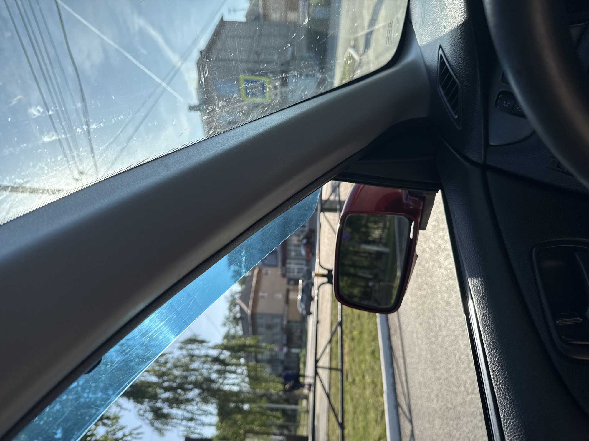 The photograph, taken from inside a vehicle, shows a partial view of the dashboard, side mirror, and windshield, with the outside world visible through the windows and mirror. The interior of the vehicle appears to be dark, with the steering wheel partially visible on the right side of the image. The side mirror, reflecting the external environment, is red. The windshield is partially covered with a blue sun visor.
Outside, a building is visible through the windshield. The side mirror shows a street with a sidewalk, trees, and possibly a building or houses in the background. The image quality is somewhat compromised by reflections and dirt on the windshield. Based on the street signs seen through the windshield, the location is near Chaikovsky, Russia.