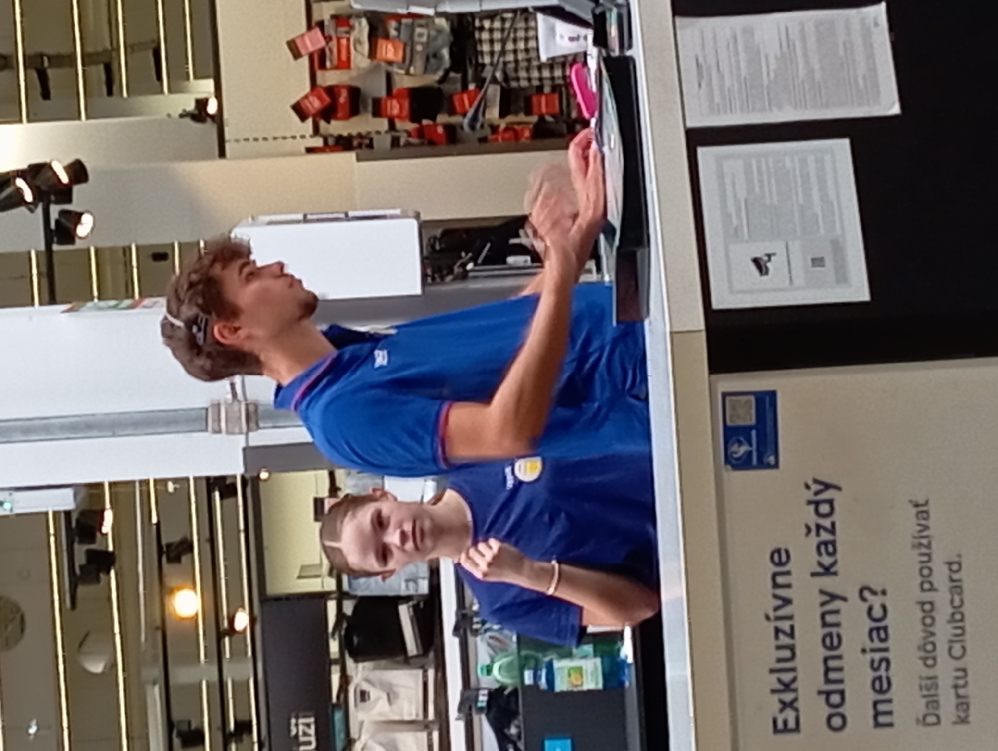 The image shows two young people, likely employees, behind a counter in what appears to be a retail store, possibly an electronics or sporting goods store, in Nitra, Slovakia. The person on the left, a young woman with dark hair pulled back, is wearing a blue polo shirt with a logo. She is looking towards the camera with a neutral expression and has her right hand raised near her face. The person on the right, a young man with curly brown hair, is wearing a blue polo shirt. He is looking down and to his right, with his hands blurred as he appears to be operating a point-of-sale system or handling merchandise. Behind them, shelves with various products are visible, along with overhead lighting and signage. In the foreground, a sign with text in Slovak, "Exkluzívne odmeny každý mesiac? Ďalší dôvod použiťvať kartu Clubcard," is visible, suggesting a loyalty program or promotion. The overall environment appears clean and well-lit.