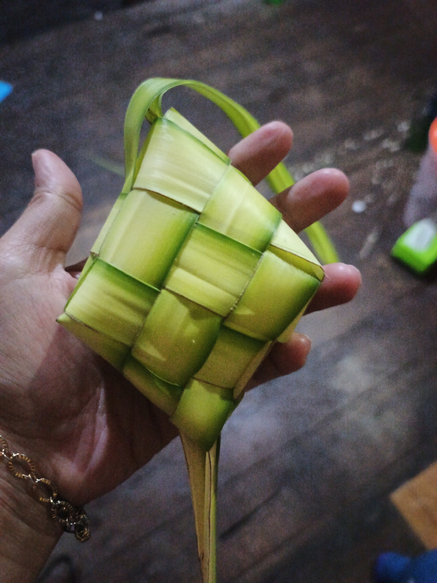 In the city of Sinar Bulan, Indonesia, a hand is holding a diamond-shaped woven palm leaf parcel, known as a "ketupat," against a blurred background of a wooden floor and a few out-of-focus objects. The ketupat is intricately woven with strips of young coconut leaves, displaying a pattern of alternating green and light yellow squares. The hand, with visible fingernails and a delicate gold chain bracelet on the wrist, is positioned to cradle the ketupat. The lighting appears to be indoor, casting a soft glow on the subject.