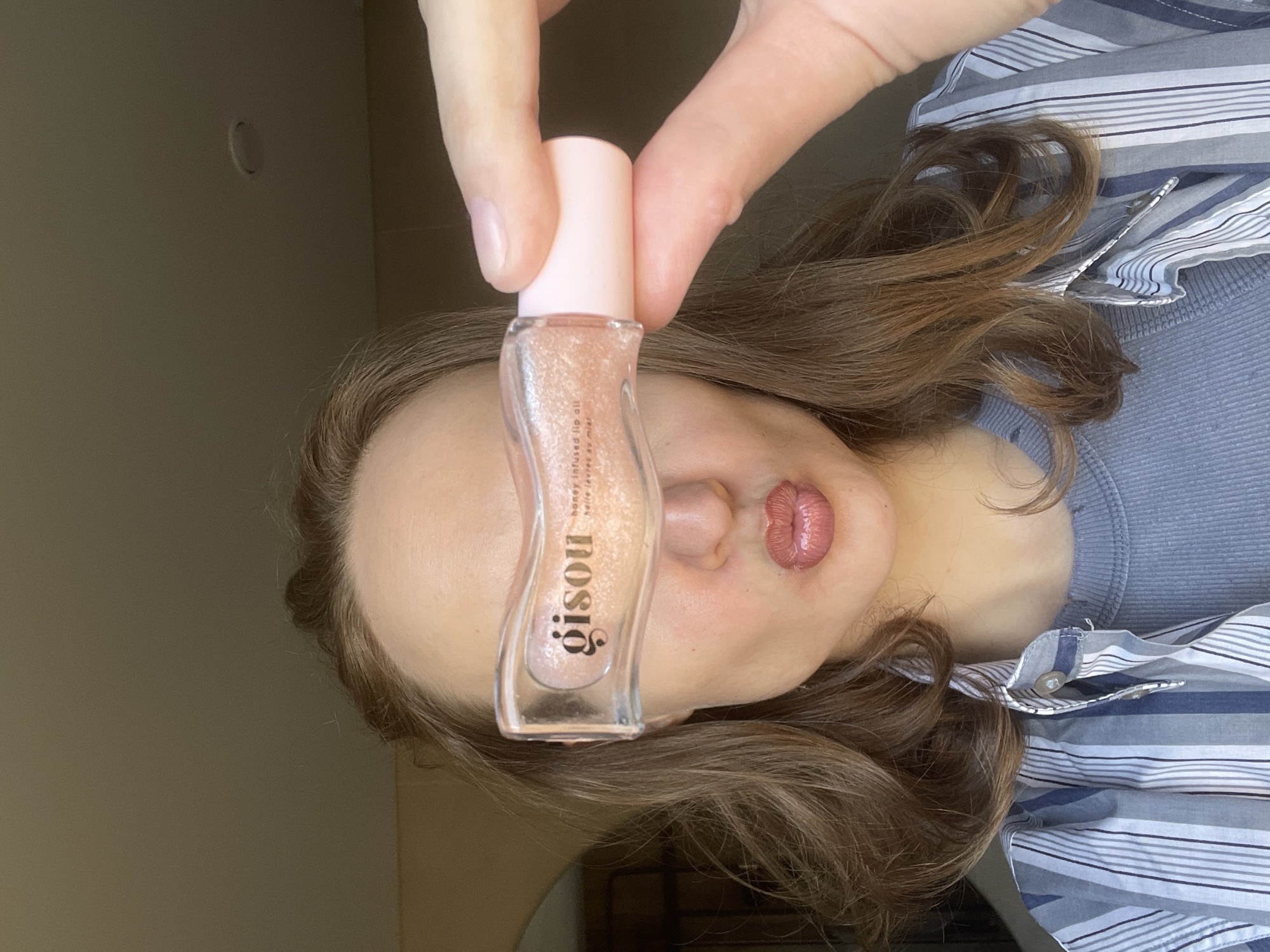 A woman is holding a clear, bottle-shaped container of lip oil in front of her face, covering her eyes and nose. The product is identified by the "Gisou" logo printed on the front. The bottle has a light pink cap. The woman has brown hair and her lips are visible beneath the lip oil. She is wearing a striped shirt and a solid grey under-shirt. The background is a plain wall. The location, Sovetsko-Nikol'skoe Lesnichestvo, Russia, is not ascertainable from the image itself.