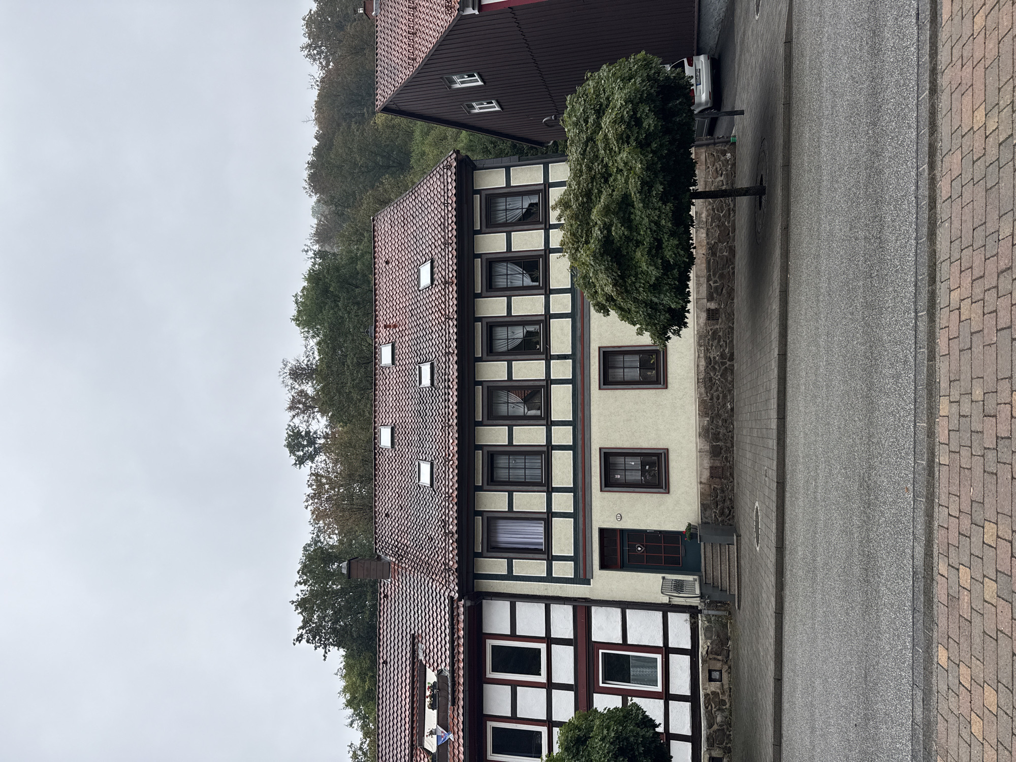 The image depicts a street scene in Walkenried, Germany, featuring a row of traditional German houses. The houses have red tiled roofs, some with dormer windows. The building on the left is white with dark red and black timber framing, and it has several rectangular windows. The house in the center has a light yellow facade with dark green and beige timber framing, and it features a dark green front door with a heart-shaped window. A set of stone steps leads up to the entrance. There are several windows on this building, some with white curtains. A manicured, bushy green tree stands in front of the central house, casting a shadow on the paved sidewalk. To the right, a dark brown, timber-clad building is visible, with a steep, sloping roof and a few white windows. A silver car is parked in the background, partially obscured by the tree and the brown building. The street in the foreground is paved with asphalt, bordered by a brick sidewalk. The sky is overcast, suggesting a cloudy day. The environment is residential and appears well-maintained. There are no people visible in the image.