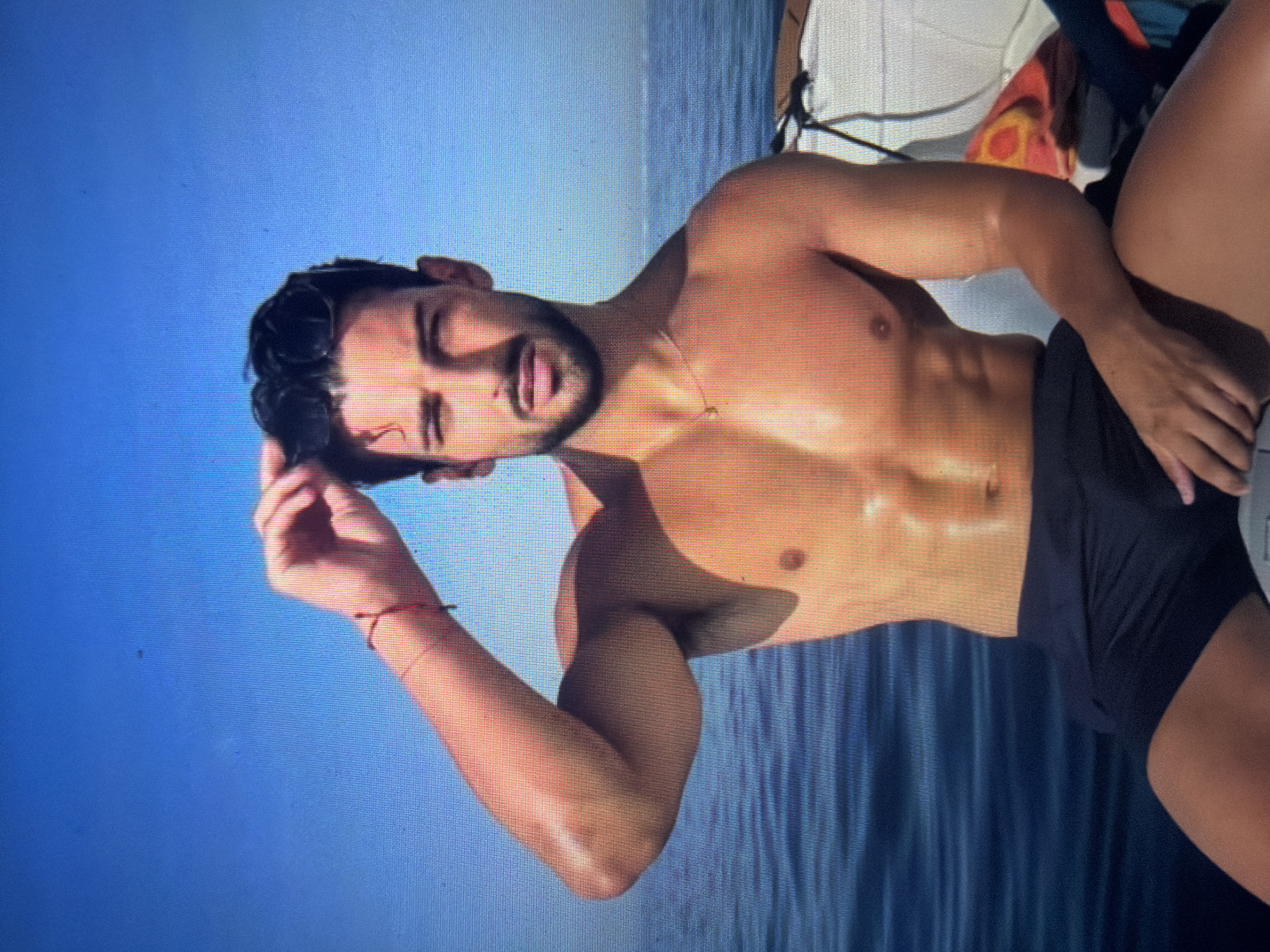 The image displays a young adult male with a muscular, tanned physique, dark hair, and a neatly trimmed beard. He has wet, dark hair styled away from his forehead and a direct, serious expression. He is wearing dark-colored swimming trunks or shorts, a thin gold chain necklace, and a red string bracelet on his left wrist. He is seated, facing forward, with his right arm raised, hand touching his hair, and his left arm bent and resting on his left leg. The background features a vast expanse of calm, dark blue water meeting a clear, bright blue sky at the horizon, indicating an open-air marine setting. Bright sunlight illuminates the scene, casting shadows on his body. Part of a white boat structure or seating is visible on the right side of the frame, with a colorful patterned item, possibly a towel, lying on it behind him. The specific location in Shumen, Bulgaria, cannot be determined from the visual information presented in the image.