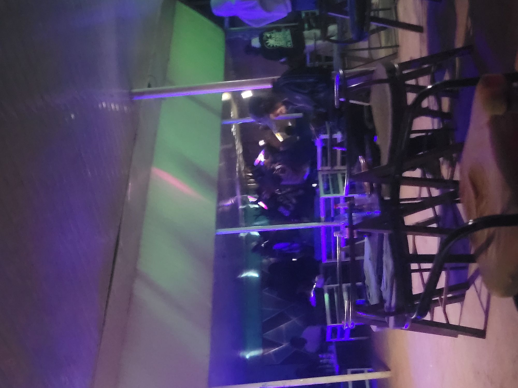 The image depicts an indoor setting, possibly a bar or lounge, with people seated at tables and chairs. The environment is illuminated by purple and green lights, creating a moody atmosphere. In the foreground, several chairs are visible, some with cushions. In the background, a group of people are gathered, some appearing to be in conversation or engaged in activities. Reflections can be seen on a mirrored surface behind the seated individuals, adding depth to the scene. The overall impression is that of a social gathering in a dimly lit establishment.