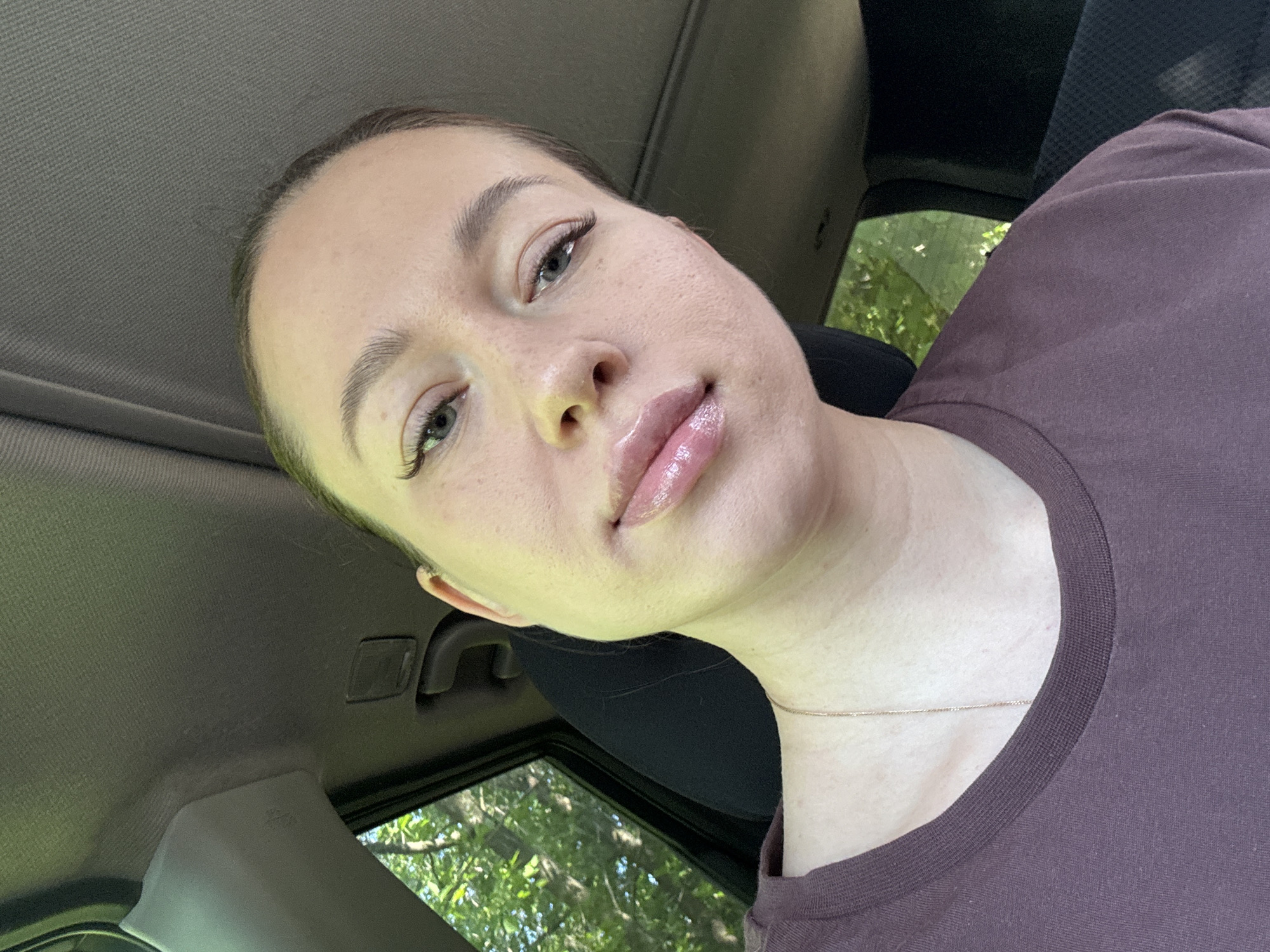 A woman is in a car. She has light skin, light eyebrows and eyelashes, and is wearing lip gloss. She is wearing a dark purple t-shirt and a delicate gold necklace. The background through the car window shows green trees, suggesting a location with vegetation. Given the location requested is in Russia and the image includes trees and a car, it's impossible to ascertain more about the specific location without further clues.