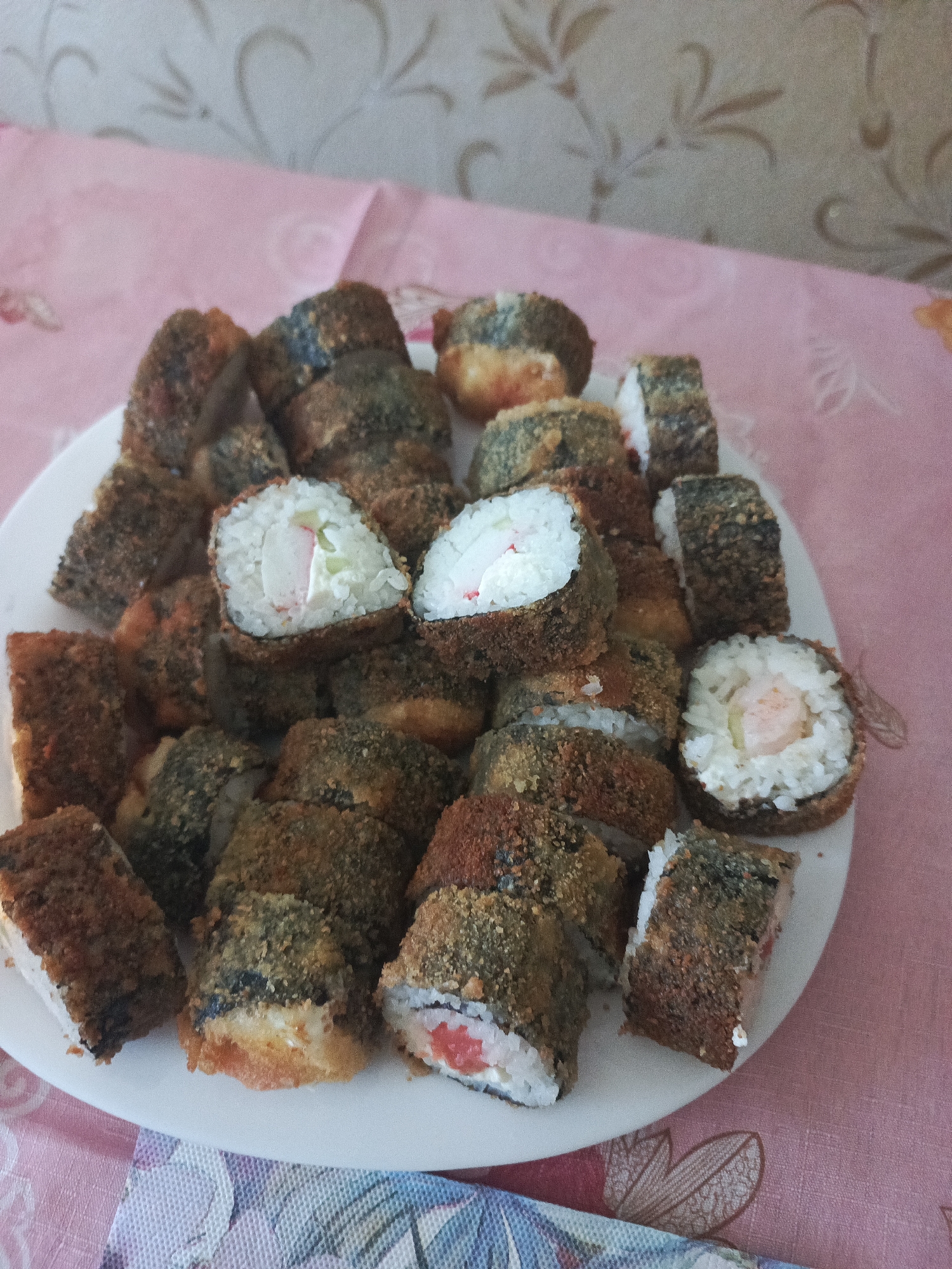 A close-up, high-angle shot shows a plate of fried sushi rolls arranged on a pink patterned tablecloth. The plate is white and filled with numerous sushi rolls, some of which are cut in half, revealing the rice and filling. The rolls are coated in a crispy, golden-brown breading. The background is a textured wall with a floral pattern. The lighting appears to be natural, possibly from a window, casting soft shadows on the food.
